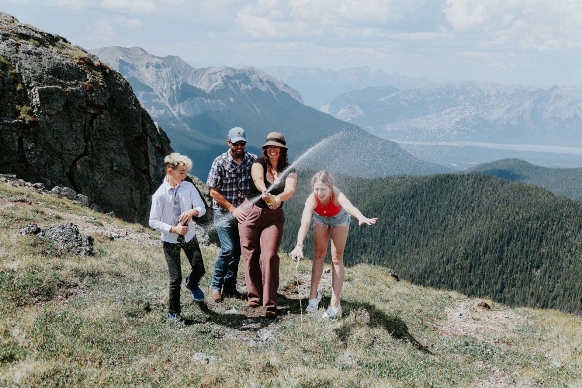 A family pops champagne on a mountain ridge with sweeping valley views.