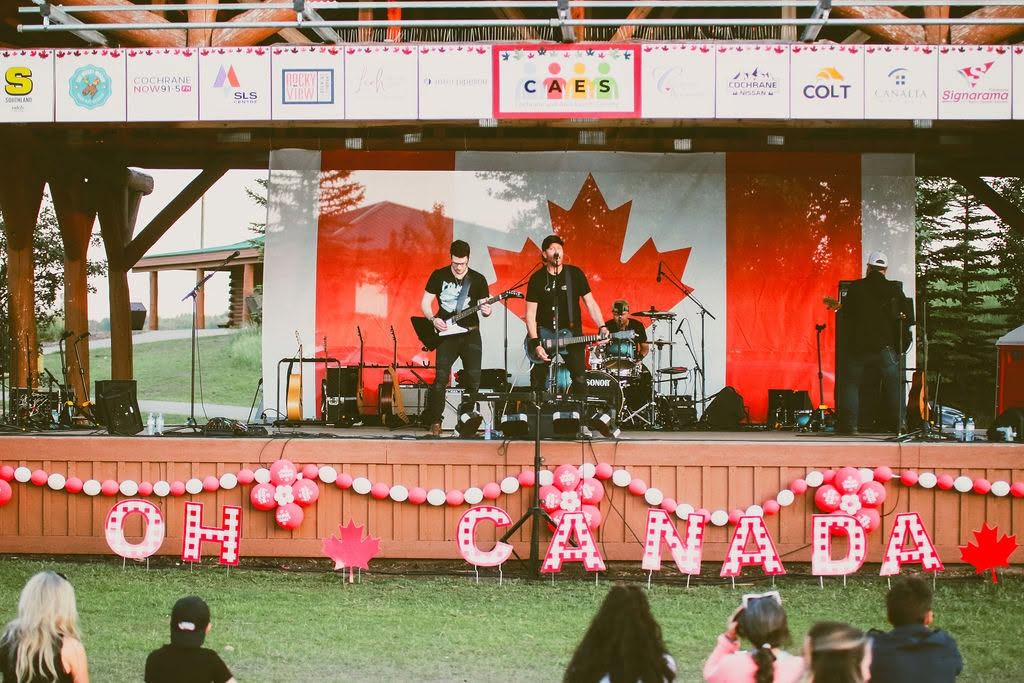 Outdoor band performs on stage with large Canadian flag and Oh Canada decorations.