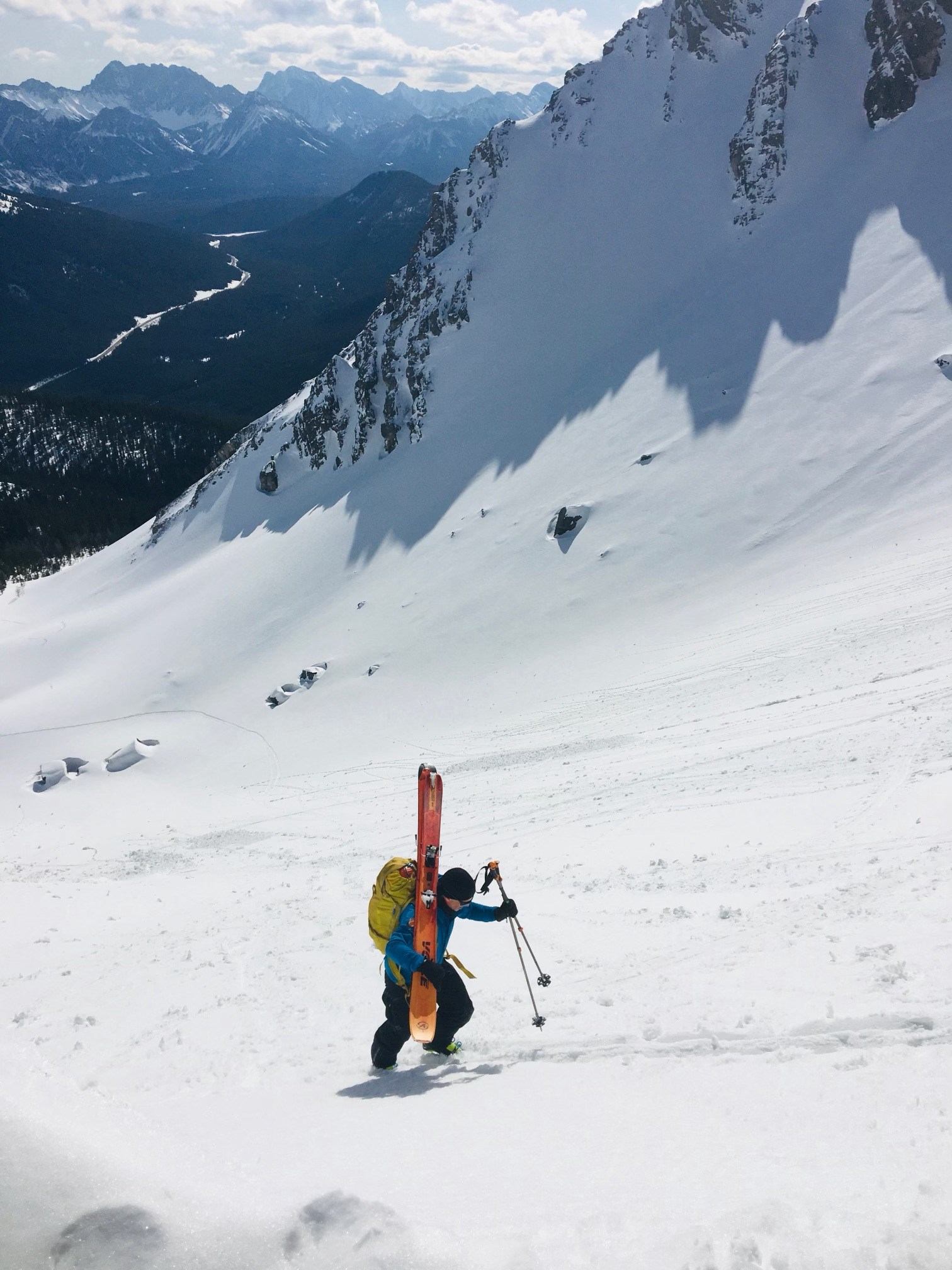 A ski mountaineer ascends a steep, snowy mountain slope with a vast mountain range and valley in the distance.