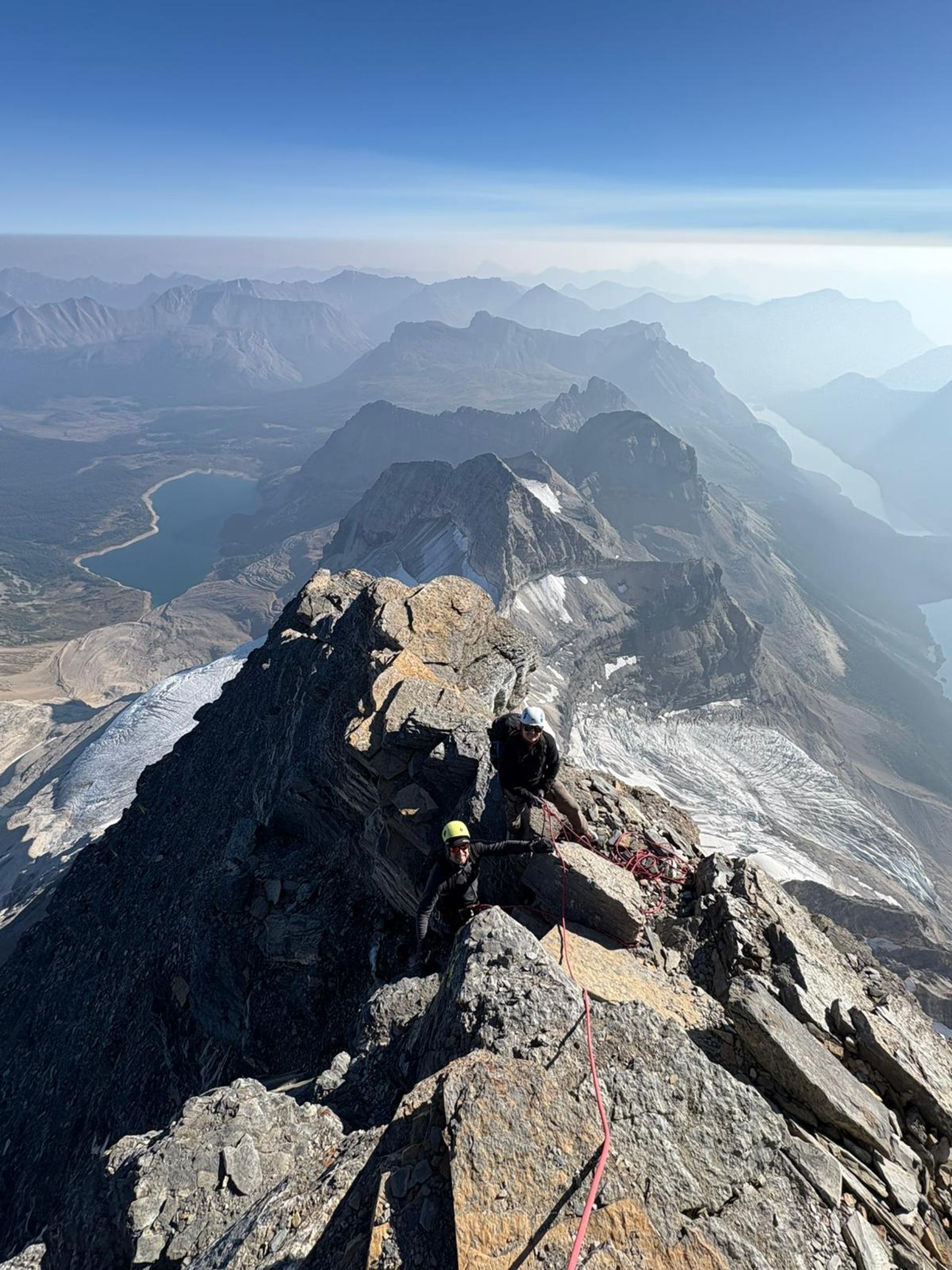 Climbers on rocky summit ridge with peaks and valley lake in the background.