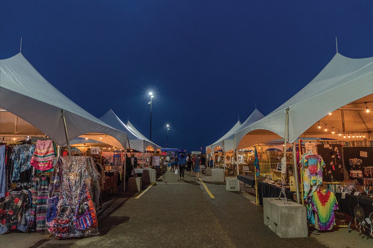Outdoor market tents at night with hanging lights, clothing racks and people walking between stalls.