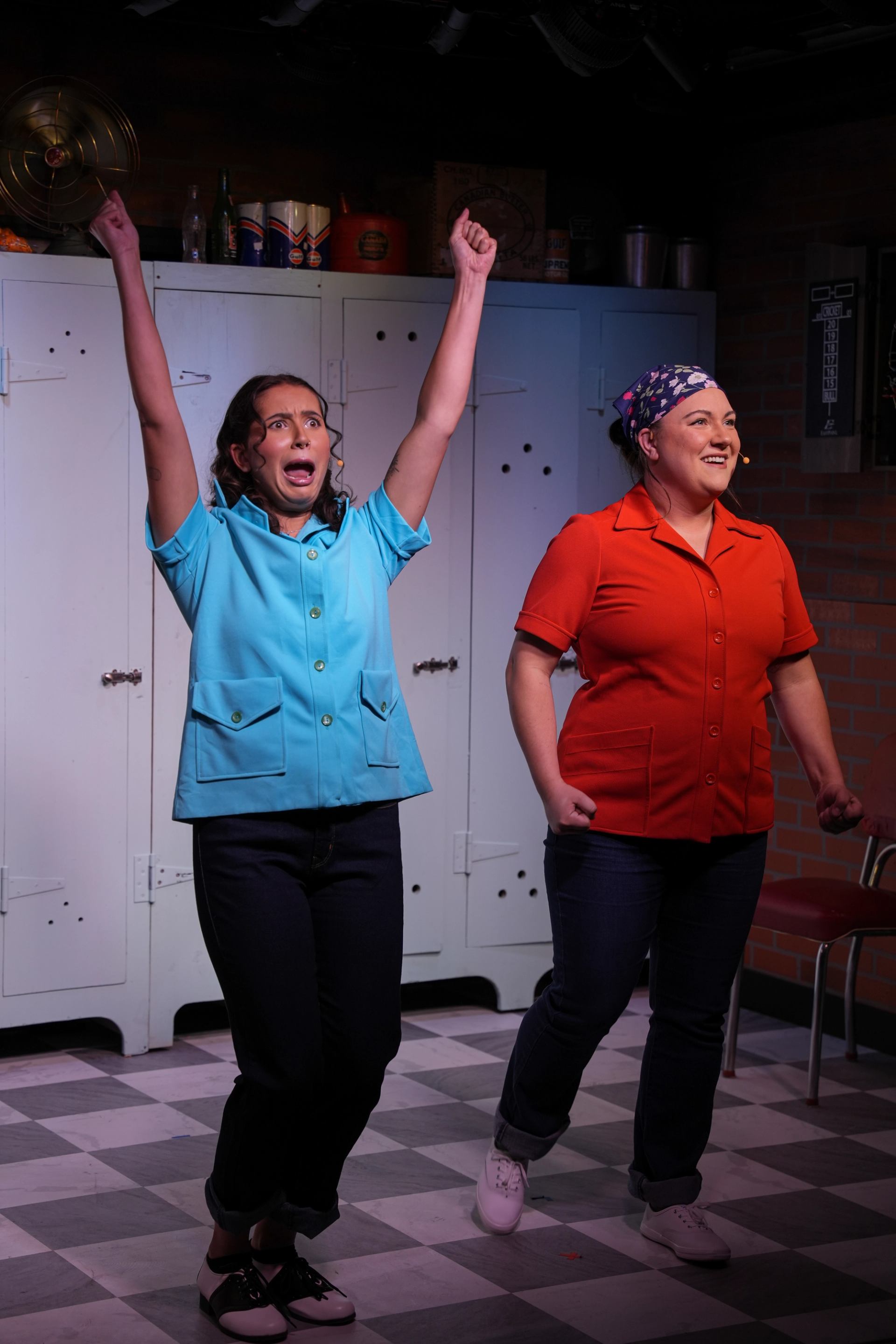 Two performers in bright uniforms sing on stage in front of white lockers.