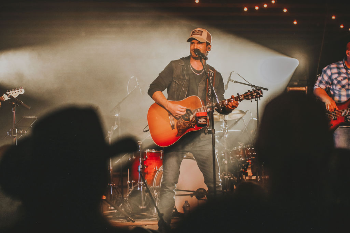 A guitar player/singer in the spotlight on stage. A few silhouettes of people in cowboy hats.
