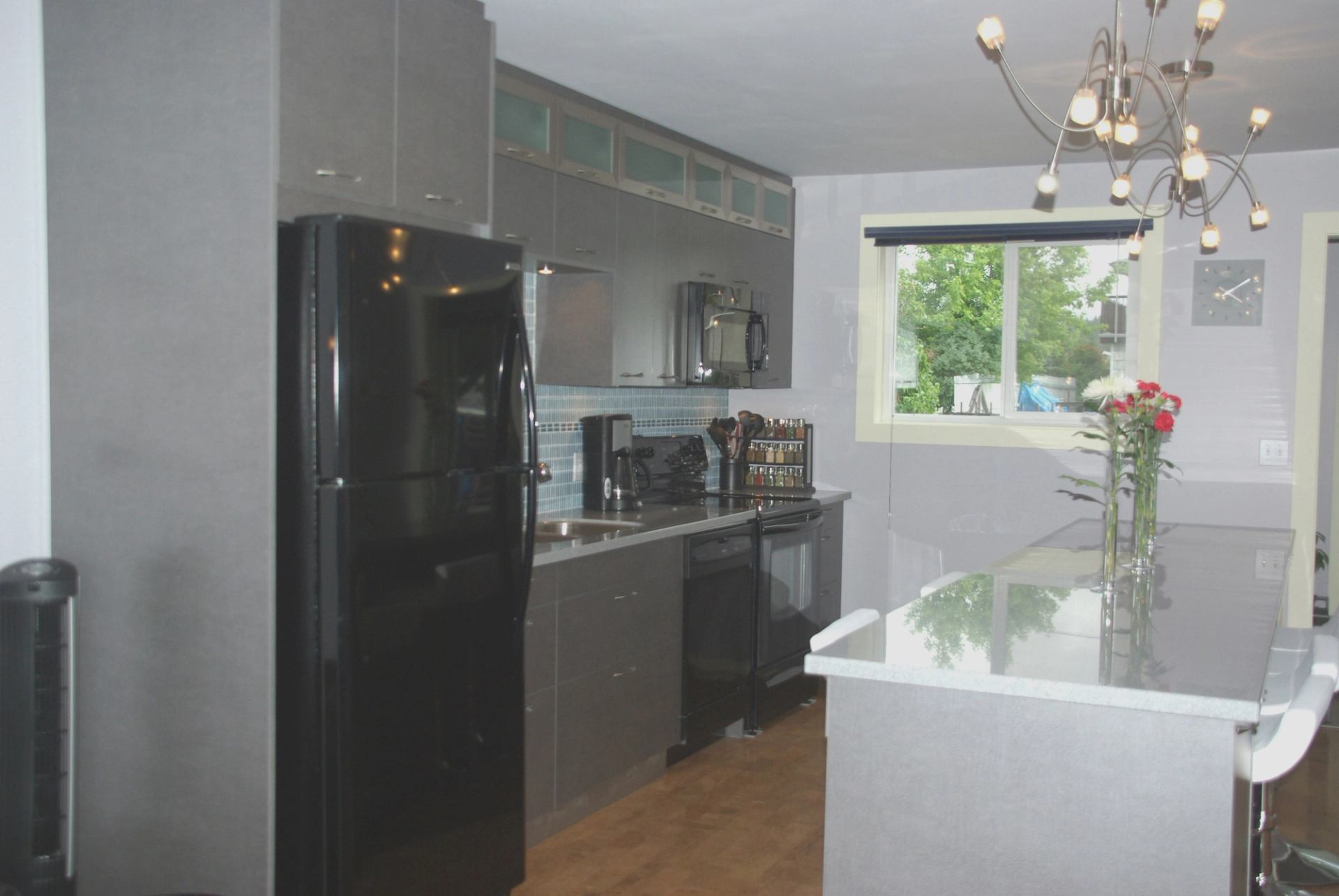 Modern kitchen with grey cabinets, black fridge, island counter, flowers, chandelier and window.