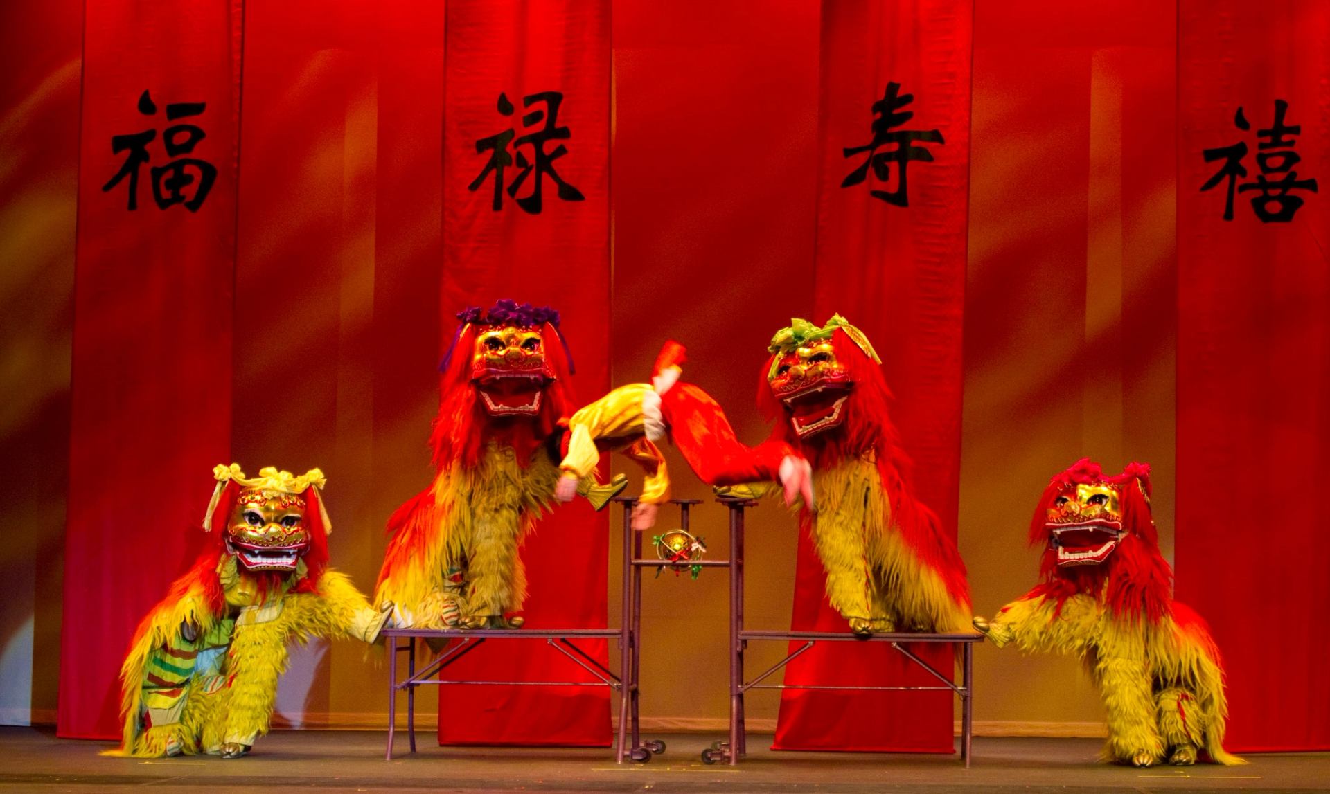 Lion dance performers in red and yellow costumes leap on platforms before red banners.