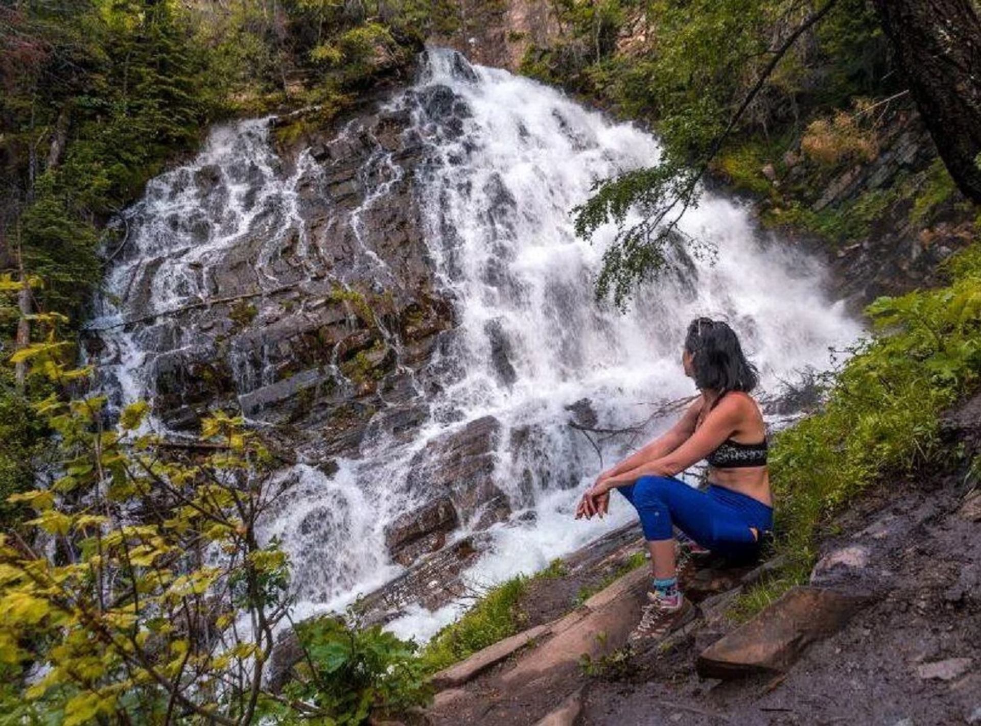 Person sitting on a rocky trail near Lower Bertha Falls surrounded by lush greenery.