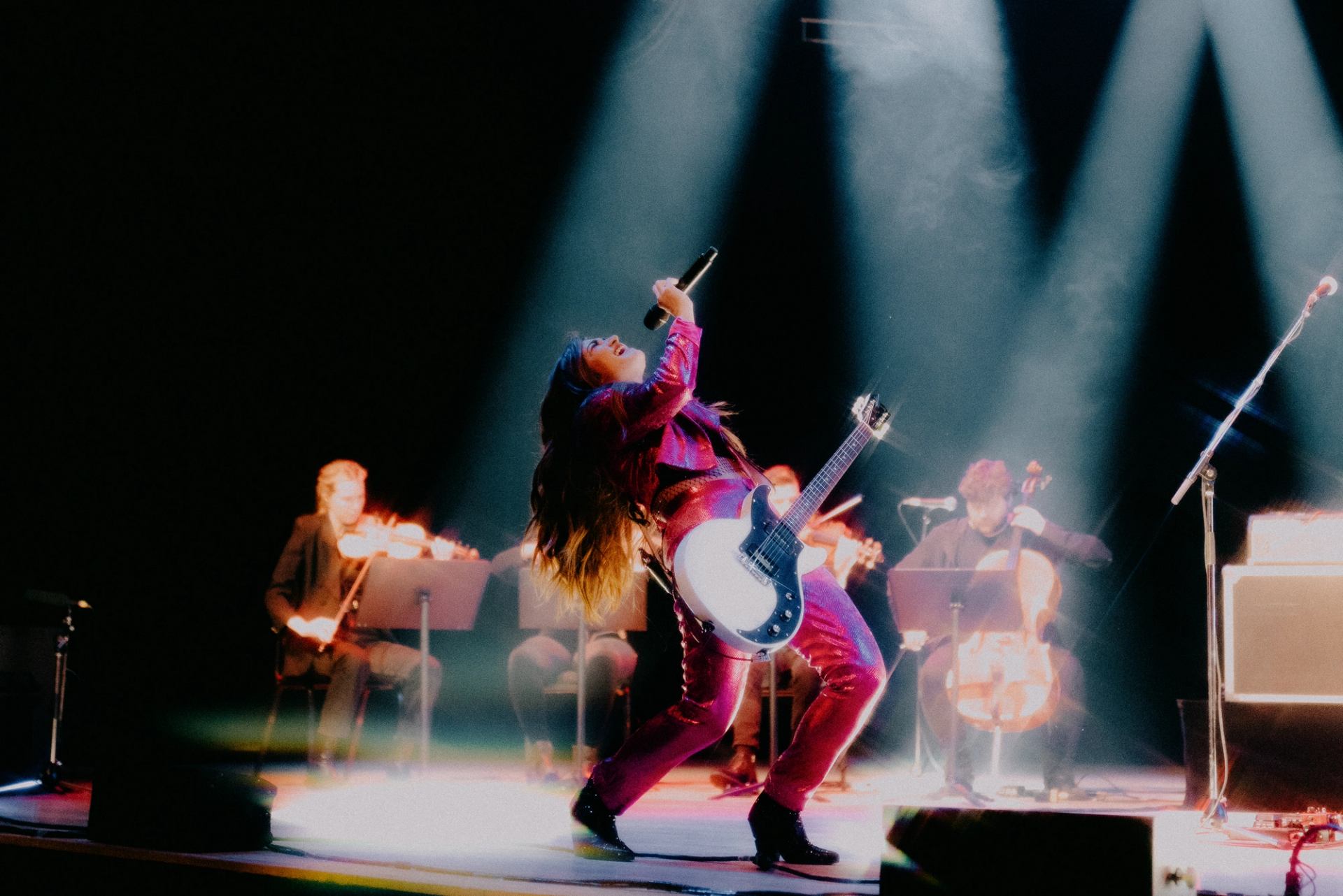 Electric guitarist in sparkly suit sings onstage with seated string musicians behind.