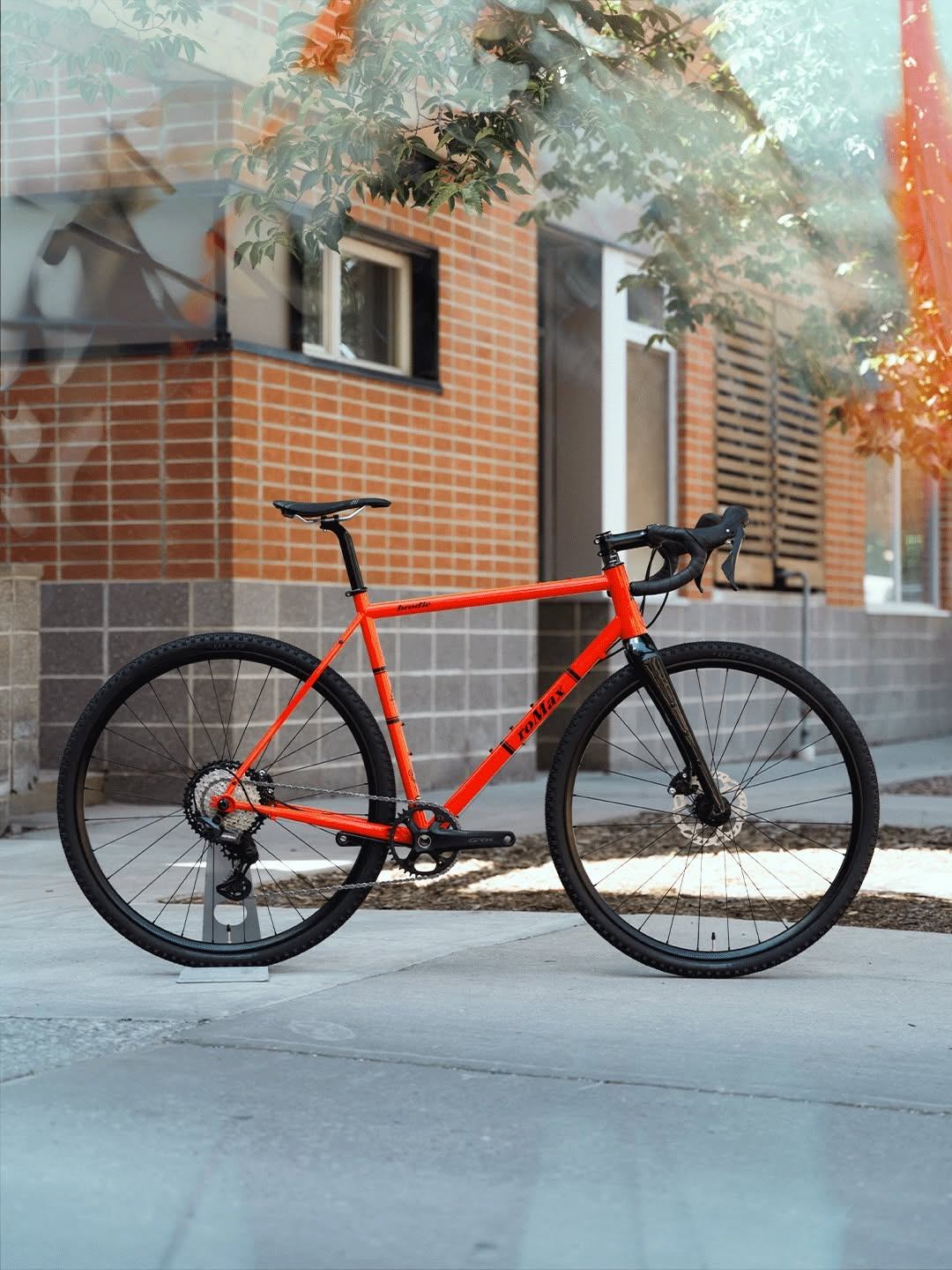 Orange bicycle with drop handlebars stands on a sidewalk beside a brick building.