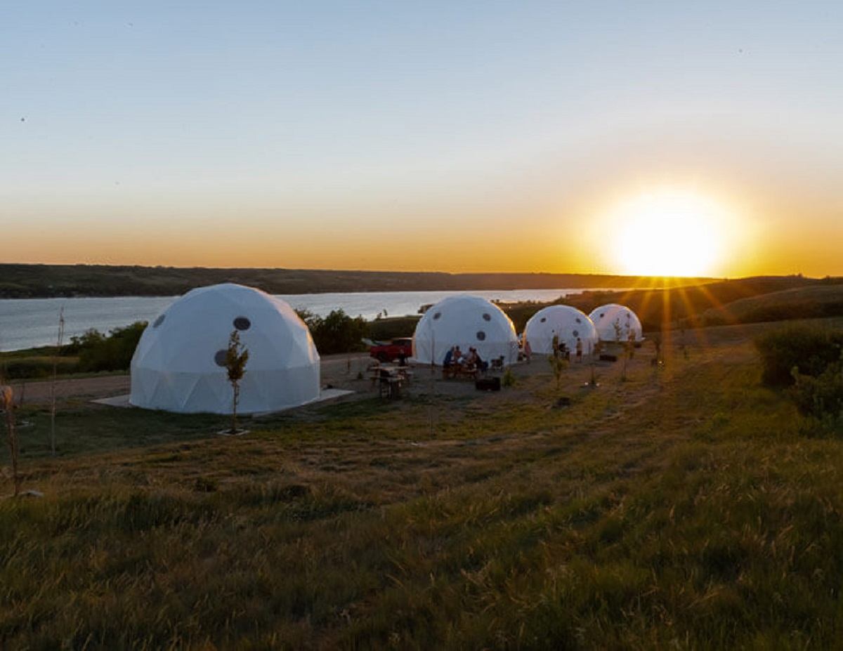 Row of white glamping domes on a grassy hill overlooking water at sunset.