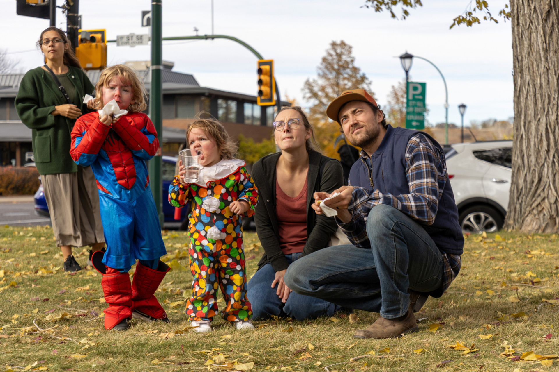 Costumed children and three adults watch something above them beside a city street.