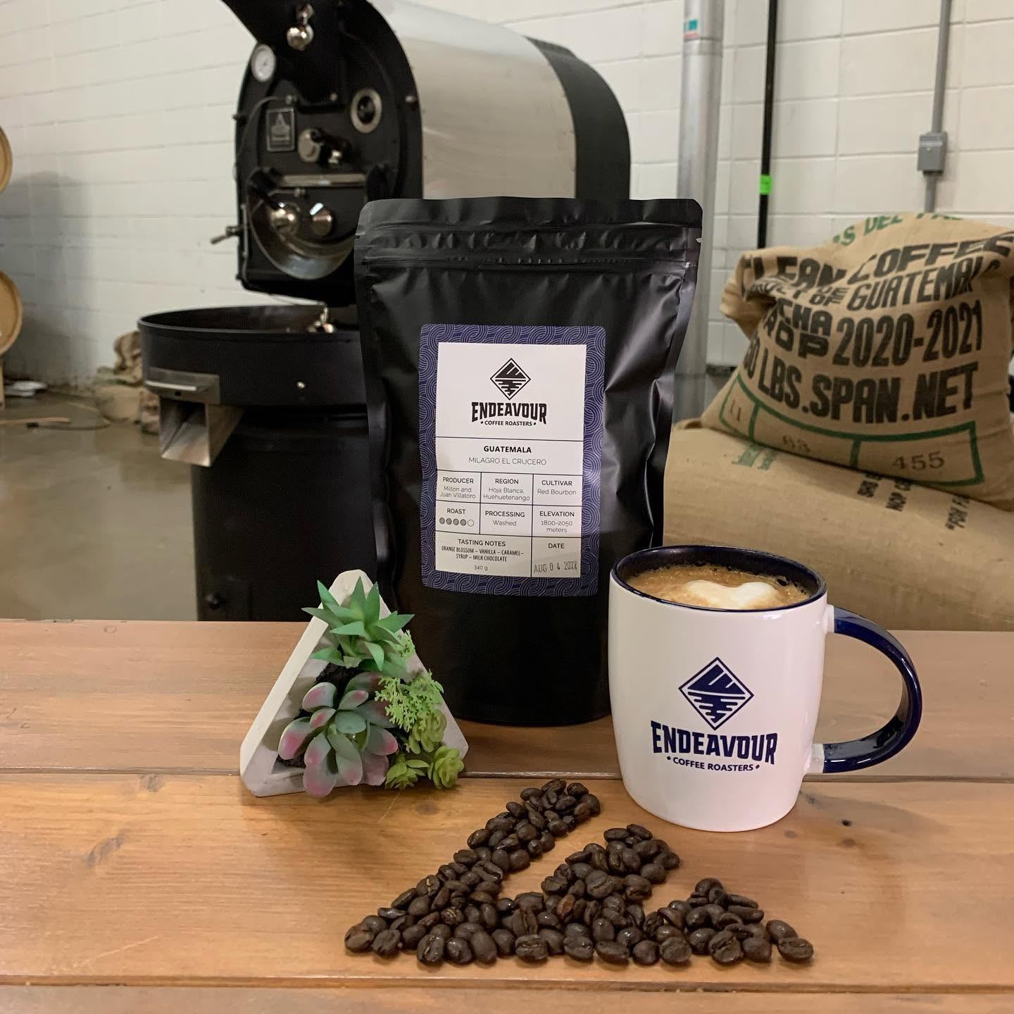 A roasted coffee bag, latte, and coffee beans sit beside roasting equipment in the roastery.