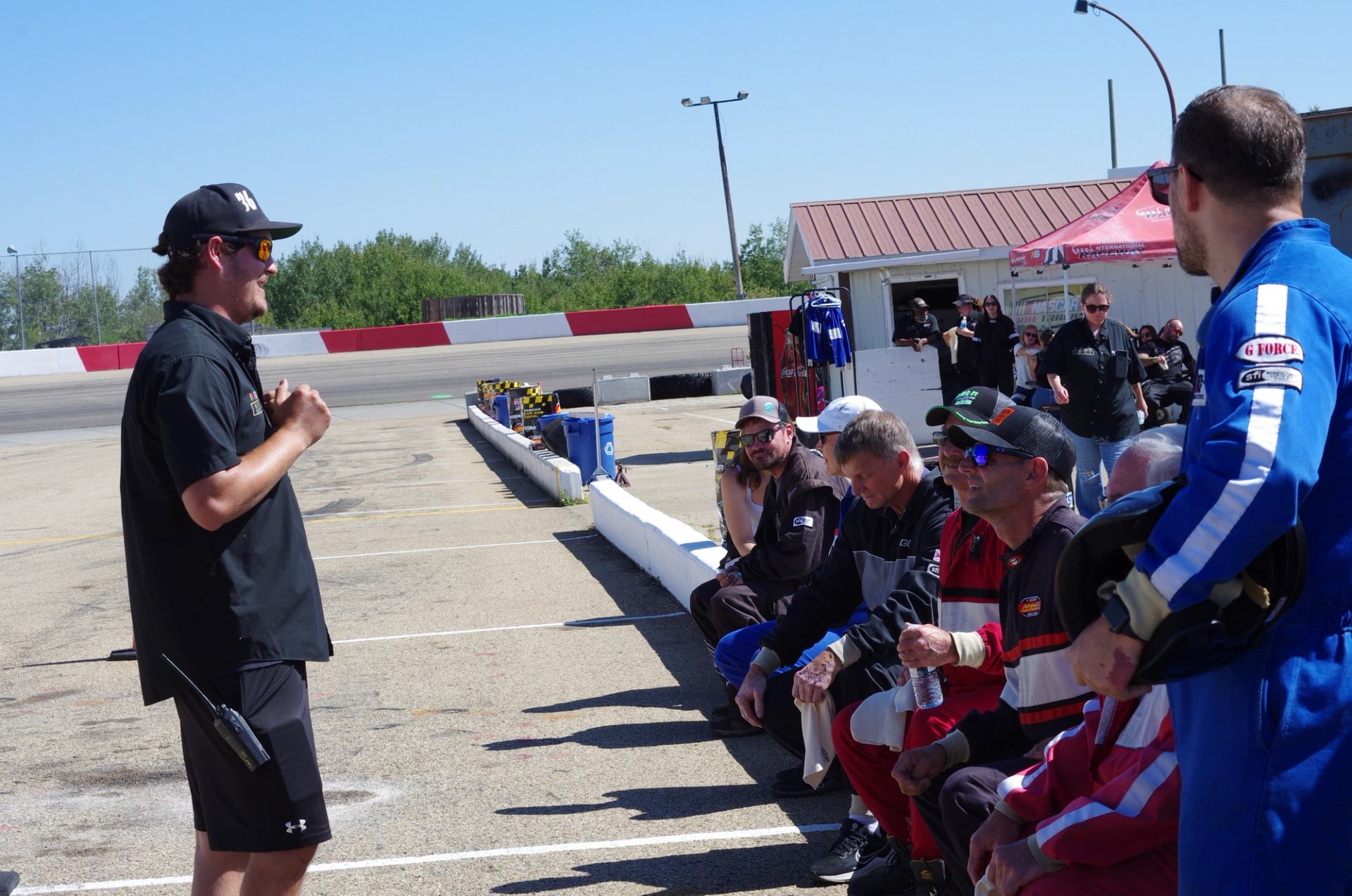 Instructor speaking to a group of people in racing suits at a racetrack.