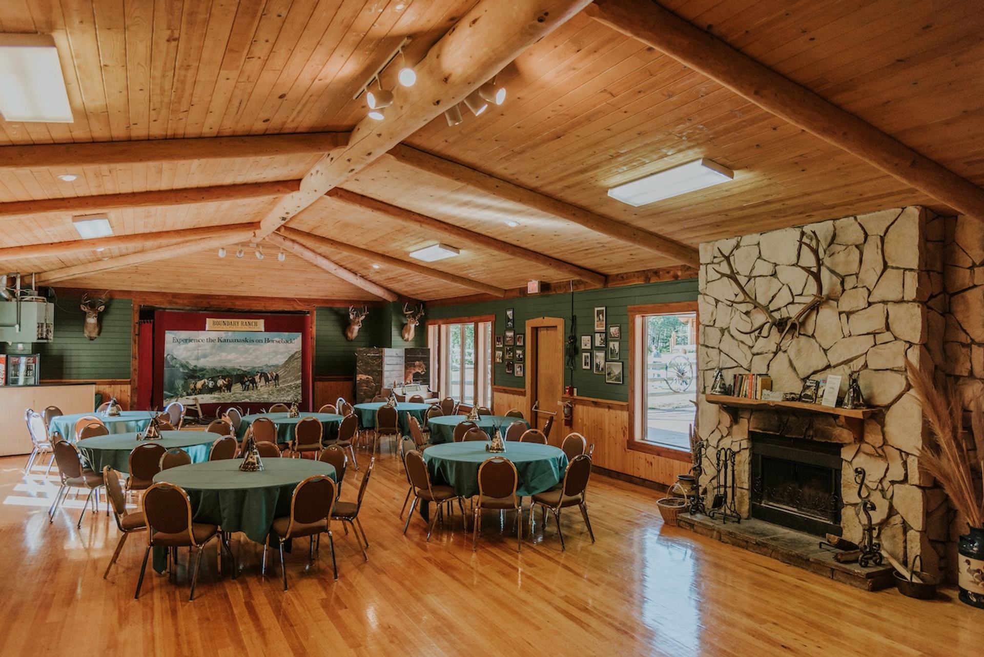 Indoor room with round tables, wood beams, large windows, and a stone fireplace.