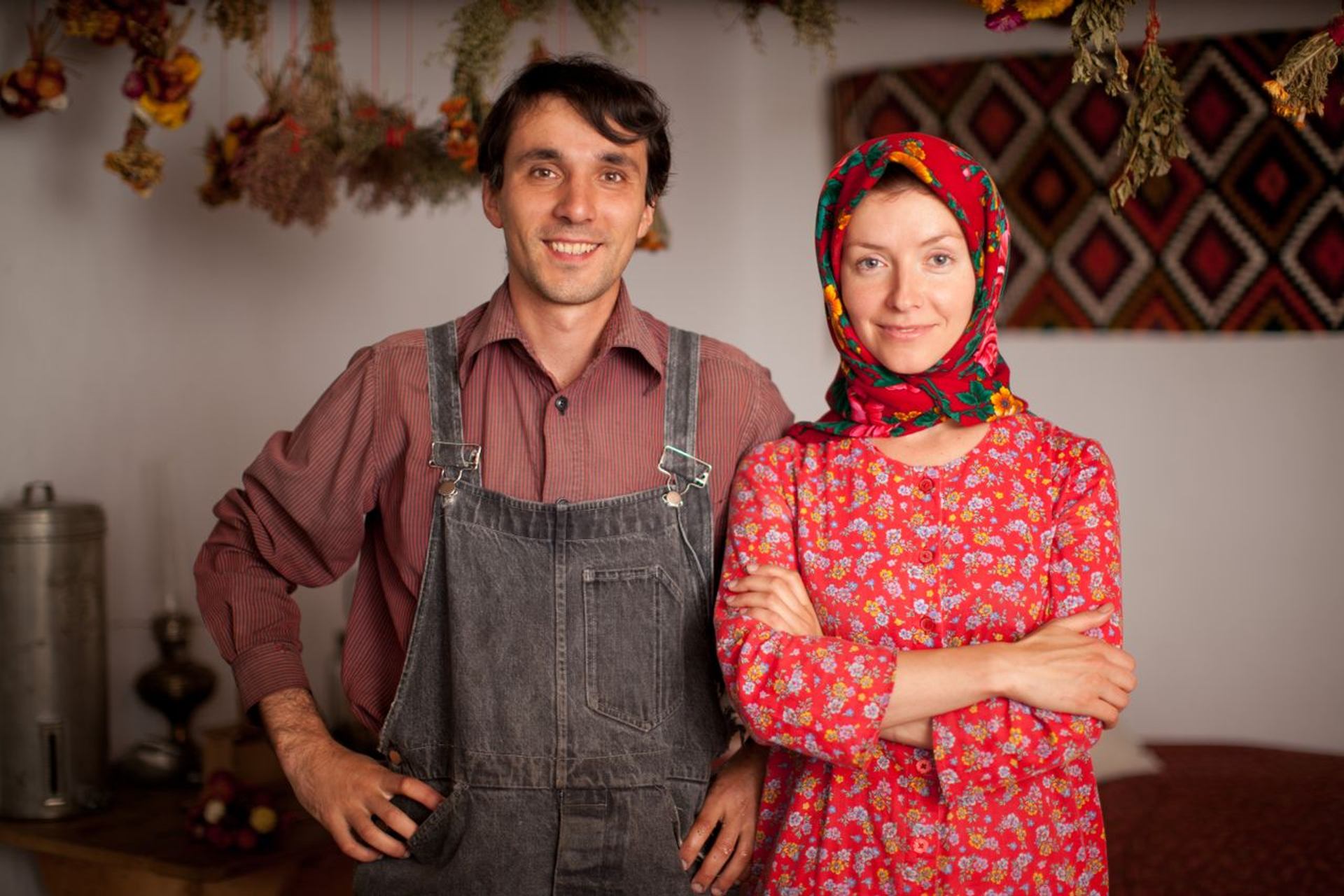 Two people in traditional clothing stand indoors with dried herbs hanging overhead.