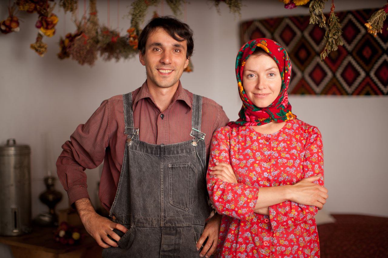 Two people in traditional clothing stand indoors with dried herbs hanging overhead.