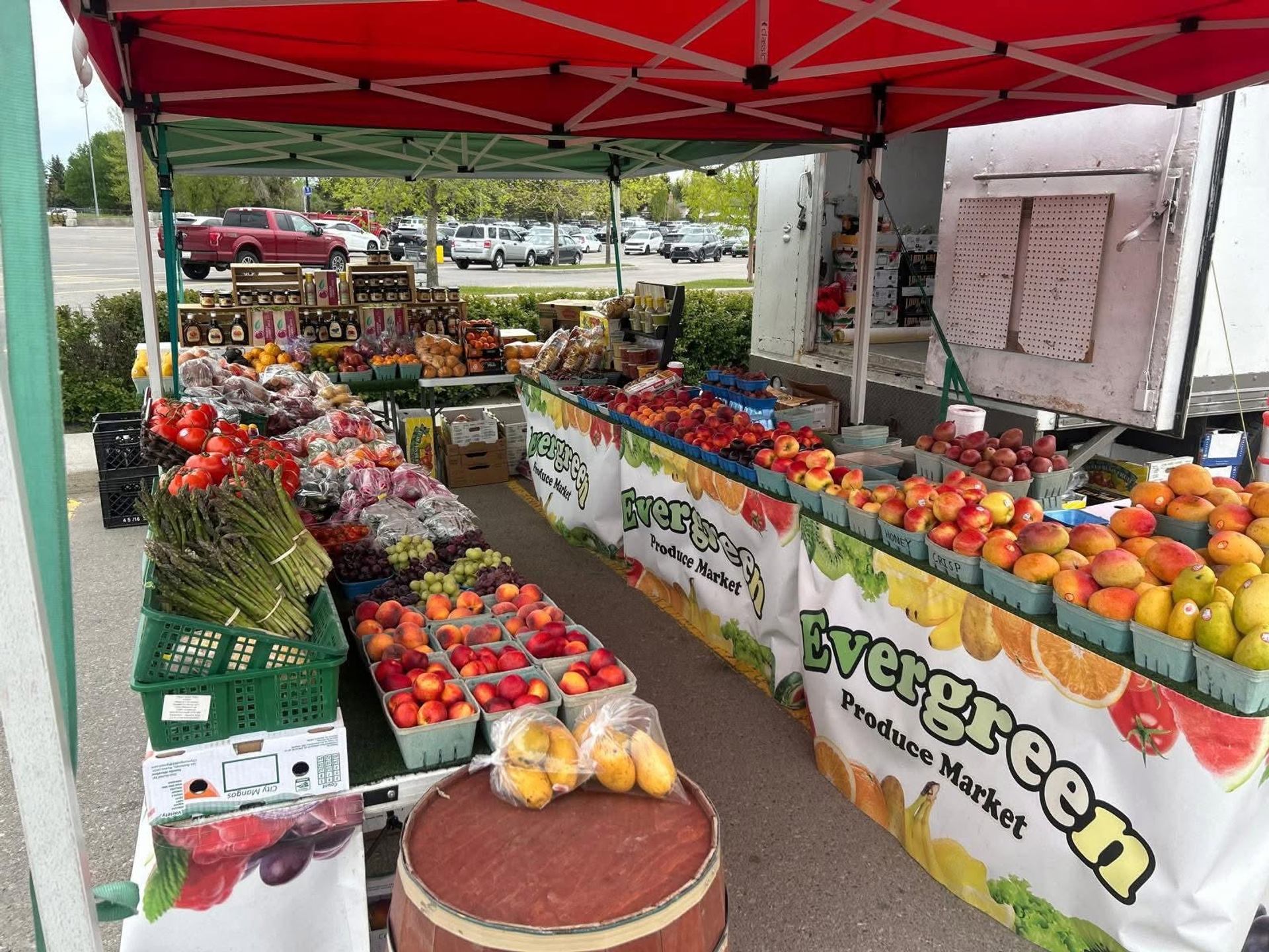 Market stall displays apples, peaches, asparagus, grapes, and assorted produce beneath canopy tents.