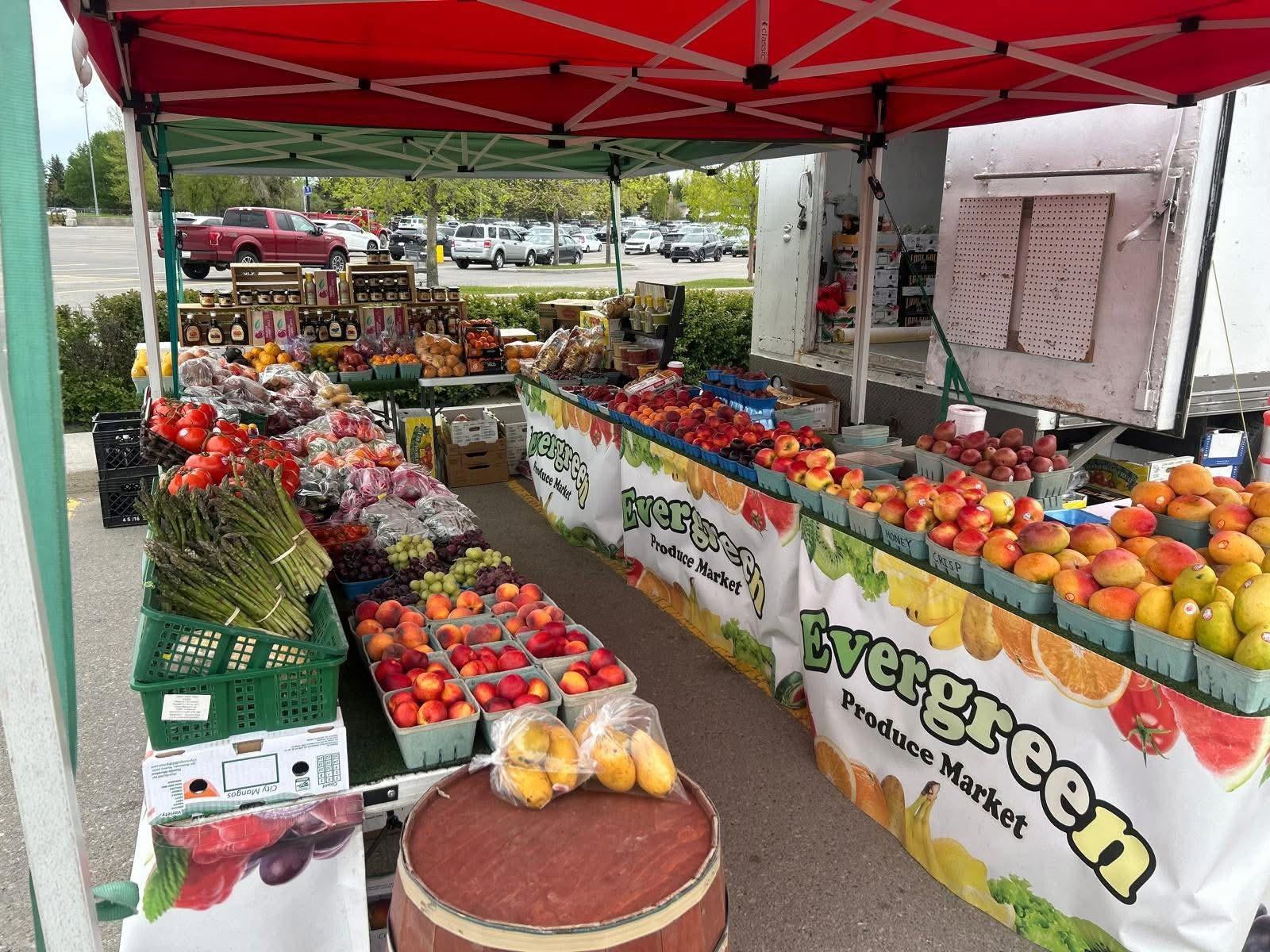 Market stall displays apples, peaches, asparagus, grapes, and assorted produce beneath canopy tents.