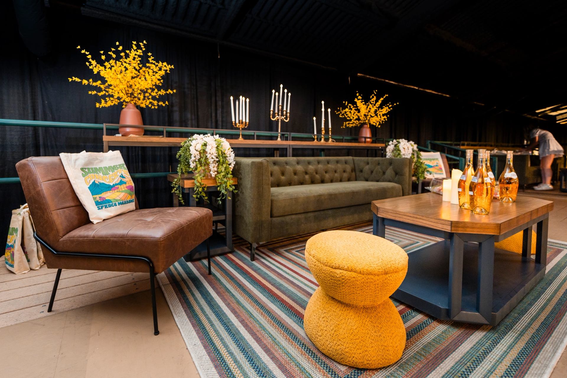 A modern lounge area with a brown armchair, green sofa, yellow stool, coffee table with bottles, and a striped rug, against a dark backdrop.