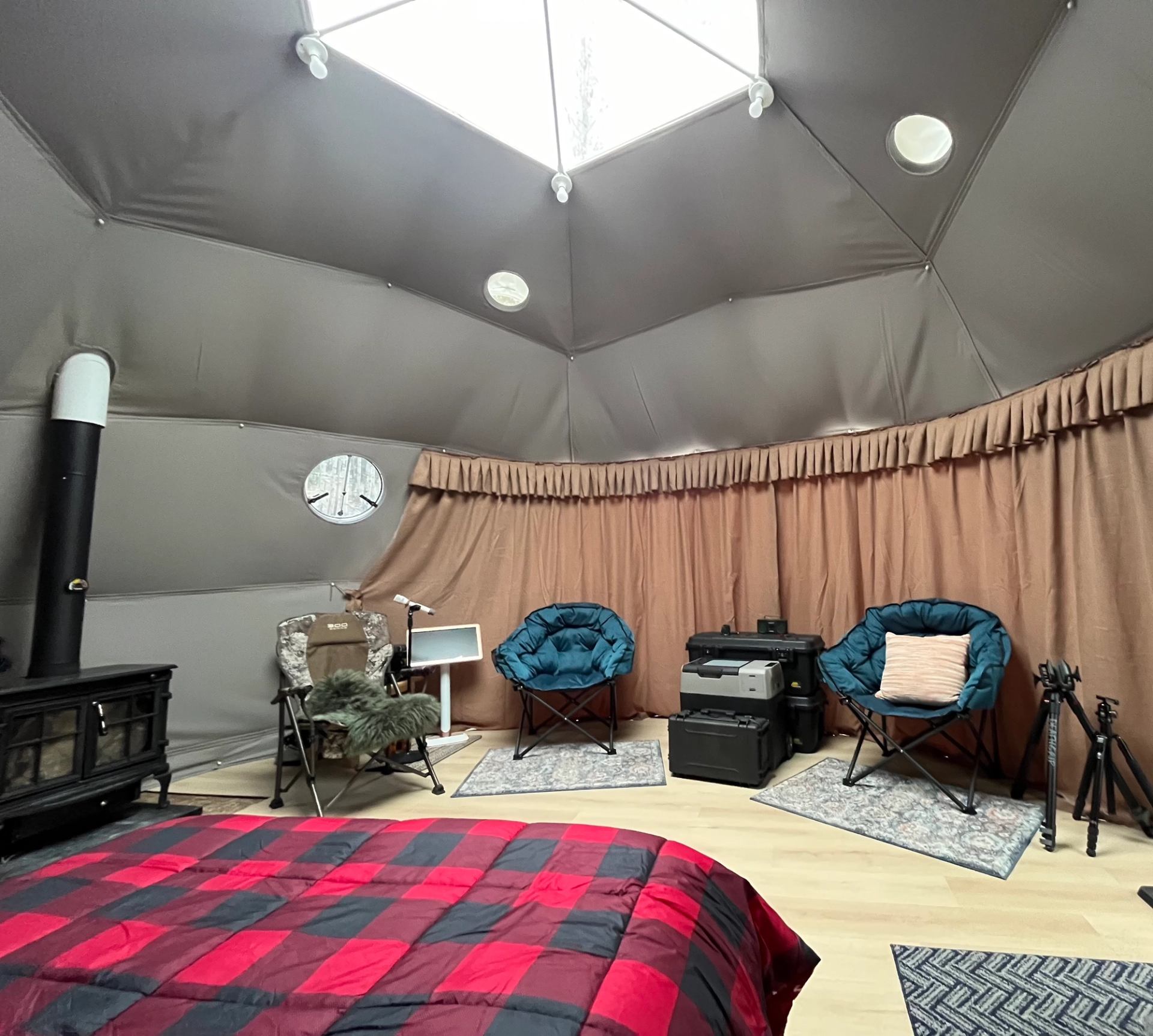 Interior of a dome cabin with chairs, wood stove, and plaid bedspread under skylight.