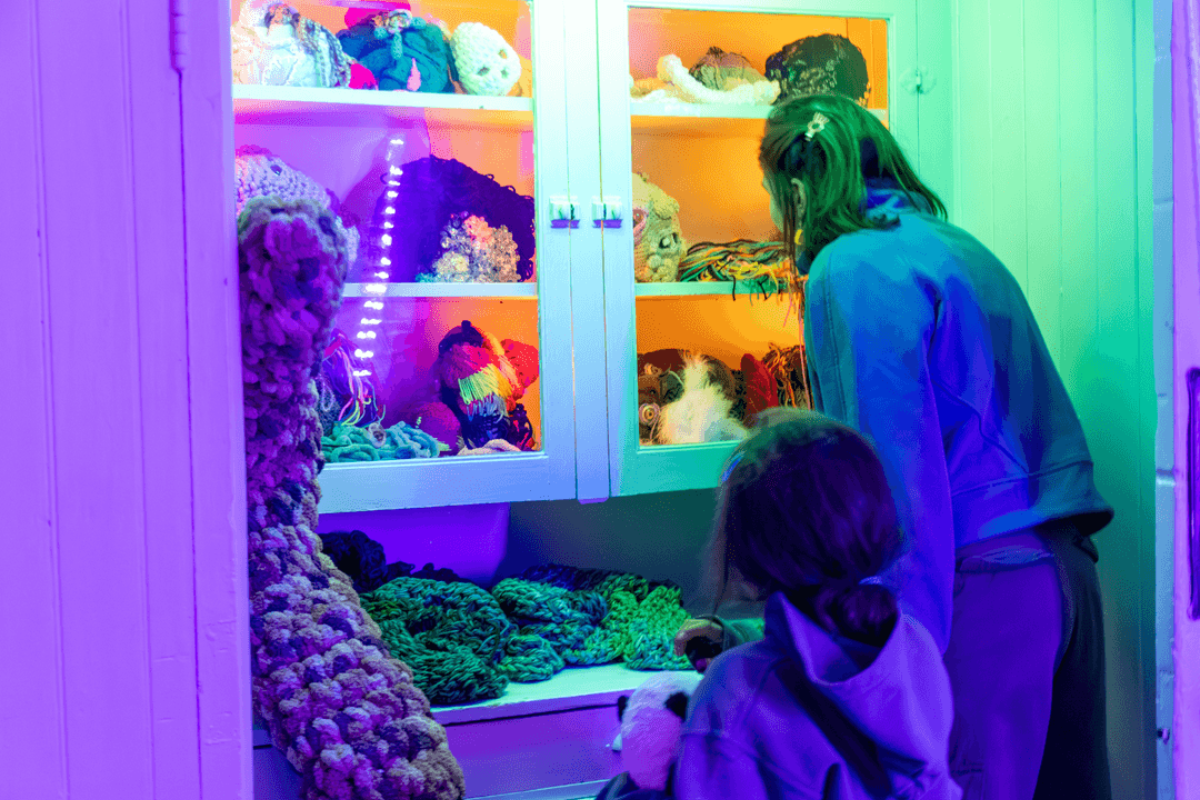 People examine colourful crocheted pieces and masks displayed inside a lit cabinet.