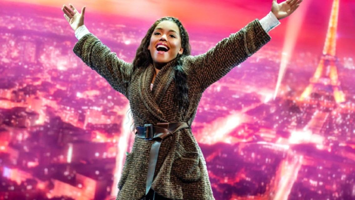 Performer on stage in a patterned coat with arms raised, singing under pink lights.