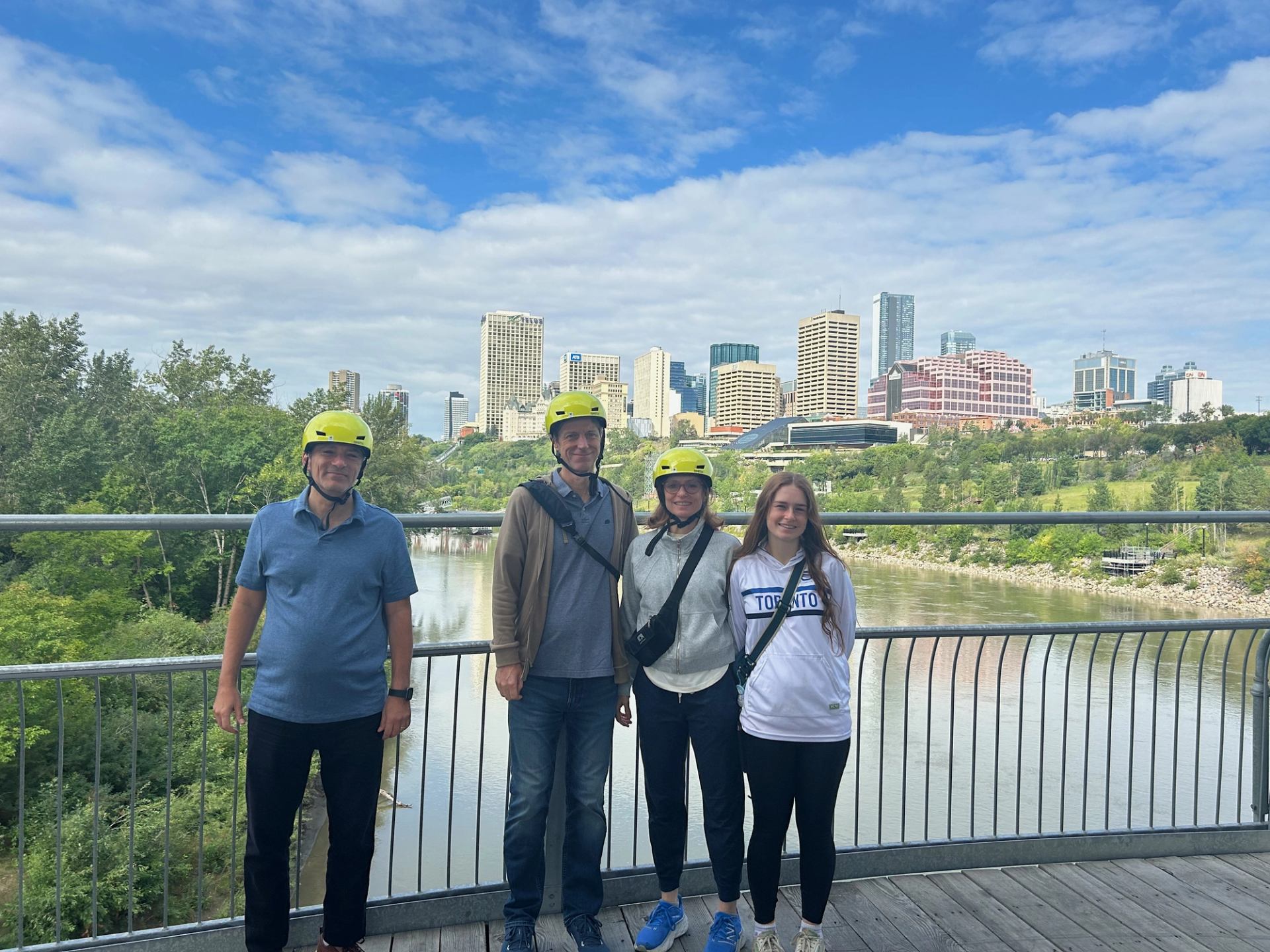Four helmeted people on a bridge with city skyline and river in the background.