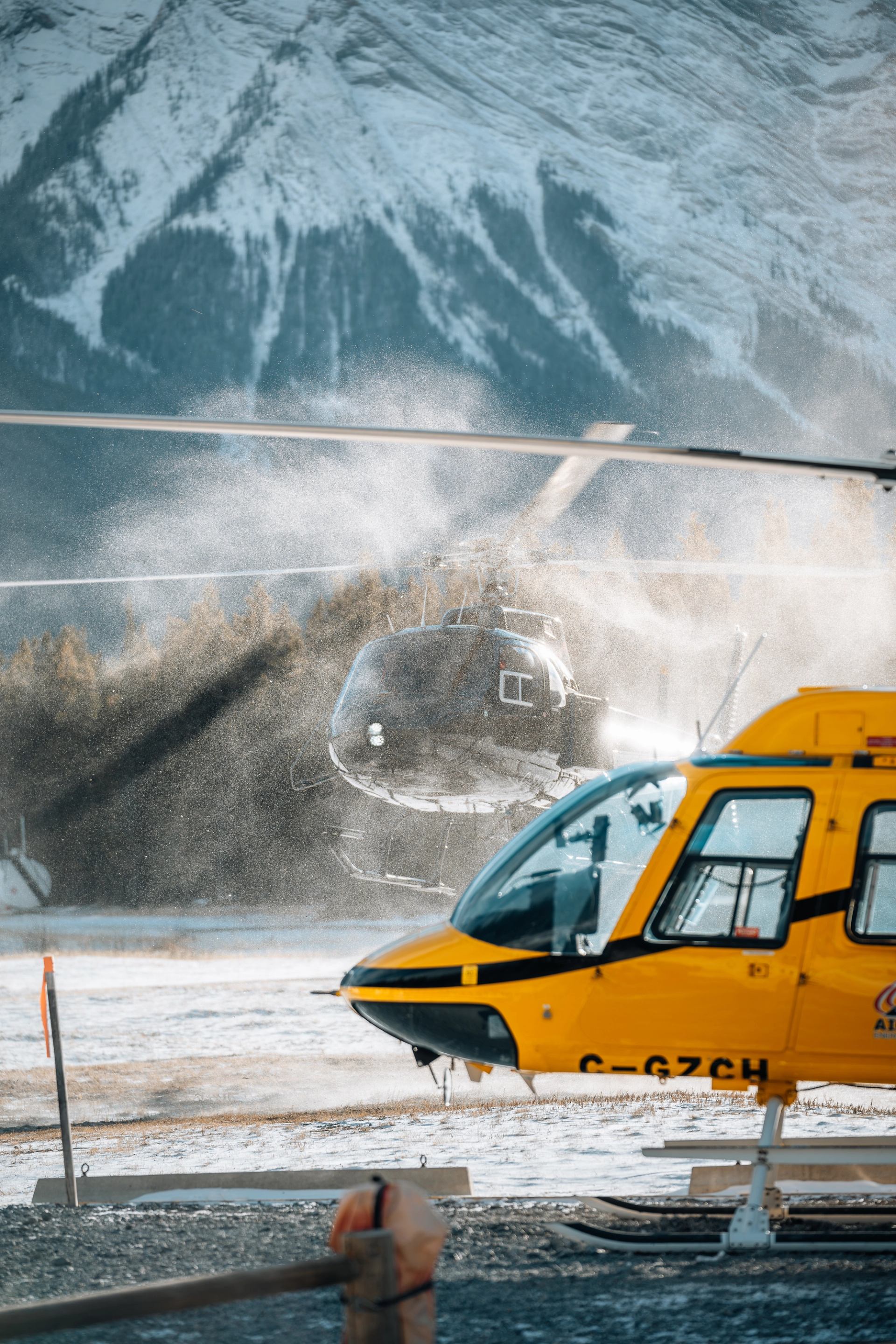 Two helicopters resting on a snowy mountain landing area in the Canadian Rockies.
