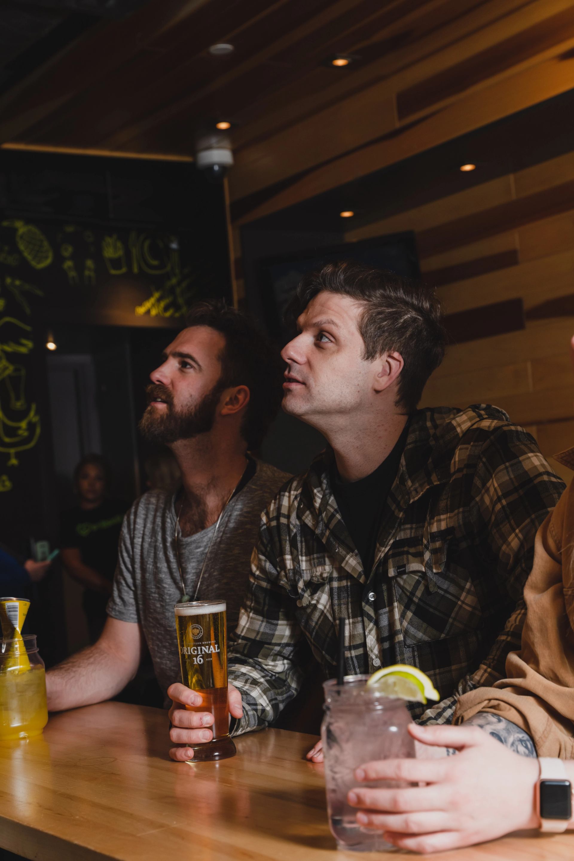 Two men at a bar looking upwards, one holding a beer, the other a cocktail.
