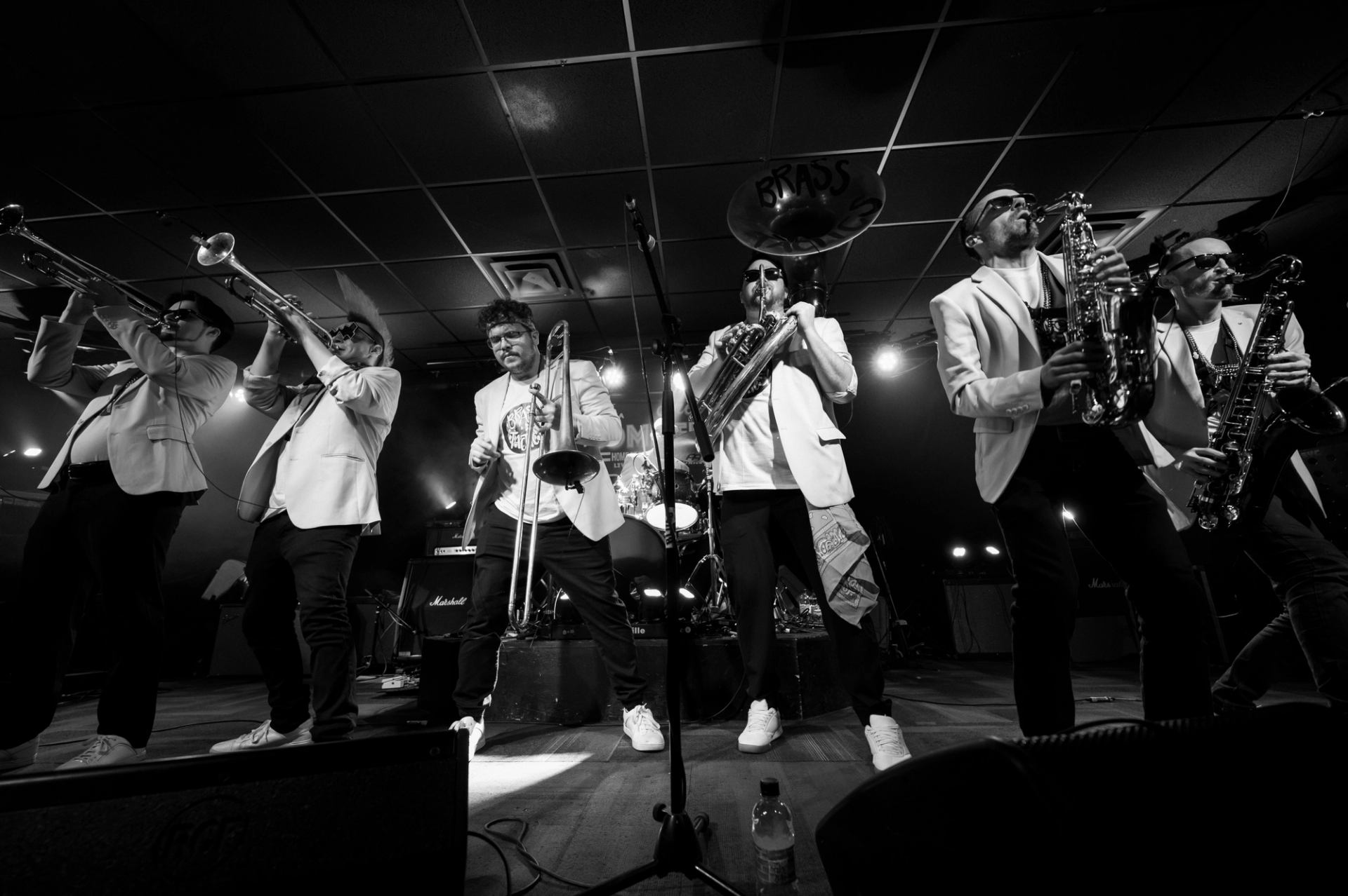 Brass band in matching jackets plays trombones, saxophones and tuba on stage under lights.