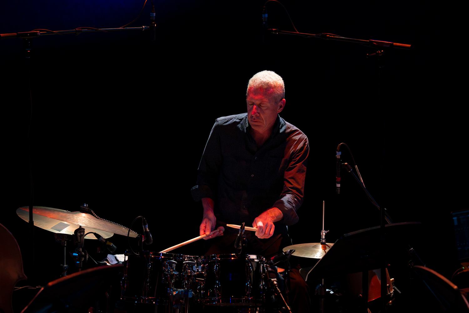 Drummer plays a drum kit on a dark stage under focused overhead lighting.