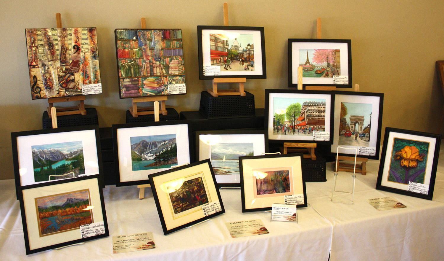 Framed landscape and city paintings displayed on easels and stands at an indoor art table.