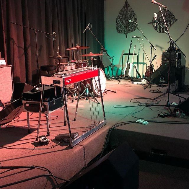 Stage with drum kit, steel guitar, microphones and guitars on stands under coloured lights.