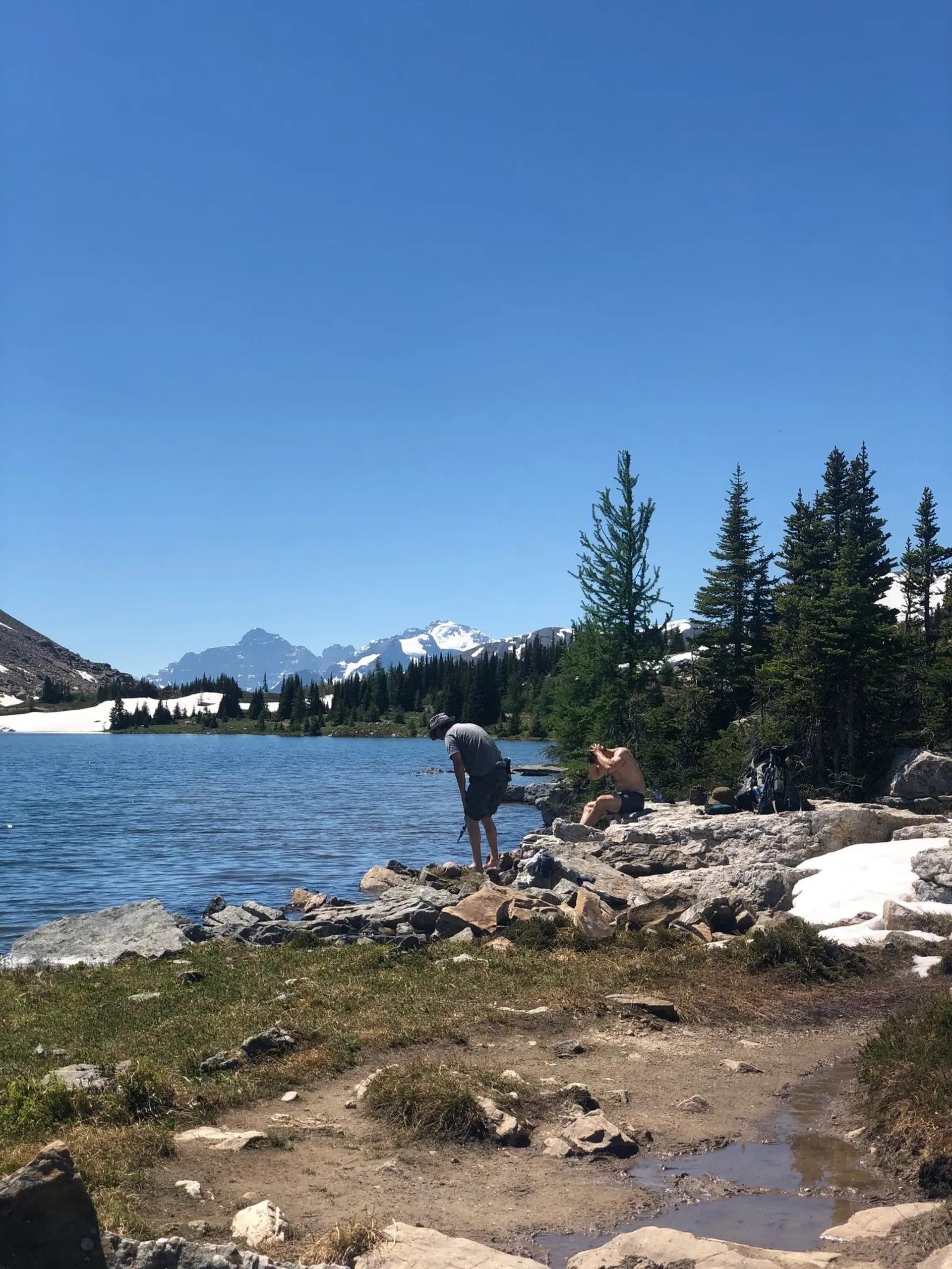 Hikers rest on a rocky lakeshore amid evergreens, snow patches and mountain peaks.