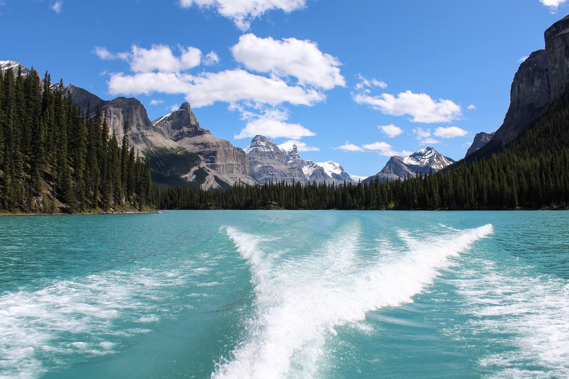 Boat wake on turquoise lake surrounded by evergreen forest and snow-capped mountain peaks.
