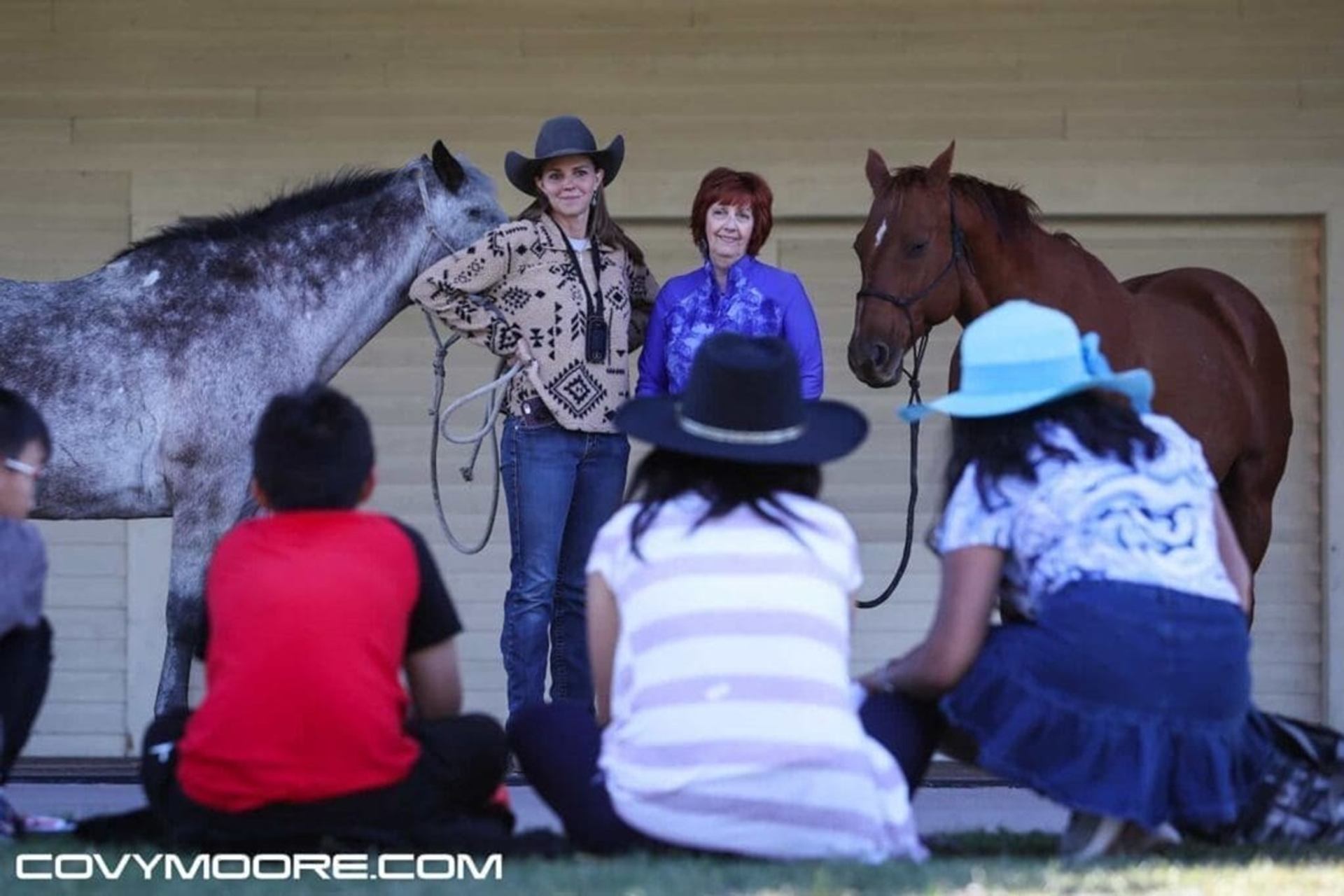 Two presenters with horses stand before seated audience wearing hats on grass.