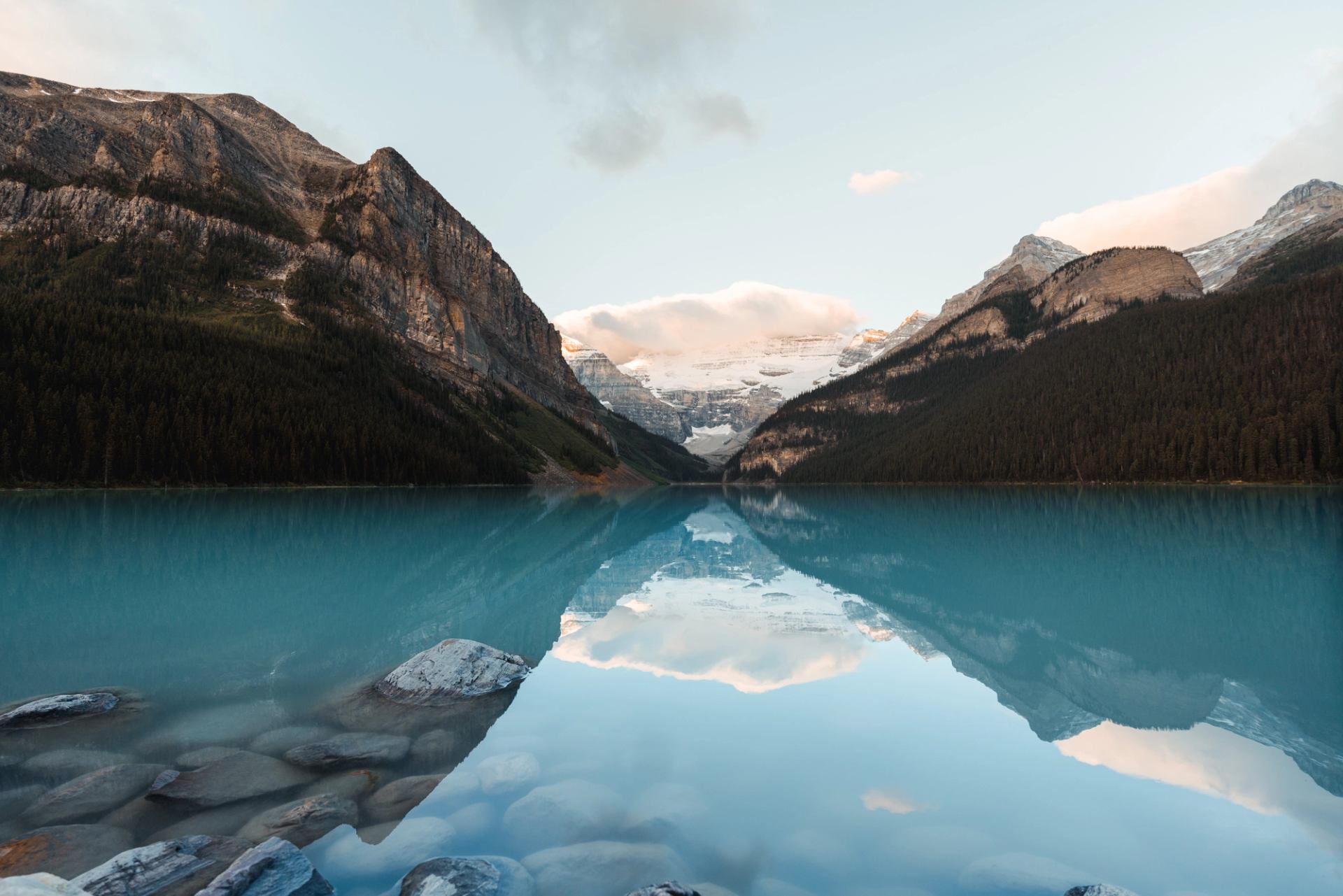 Turquoise lake reflects forested mountains, snowy peaks and clouds.