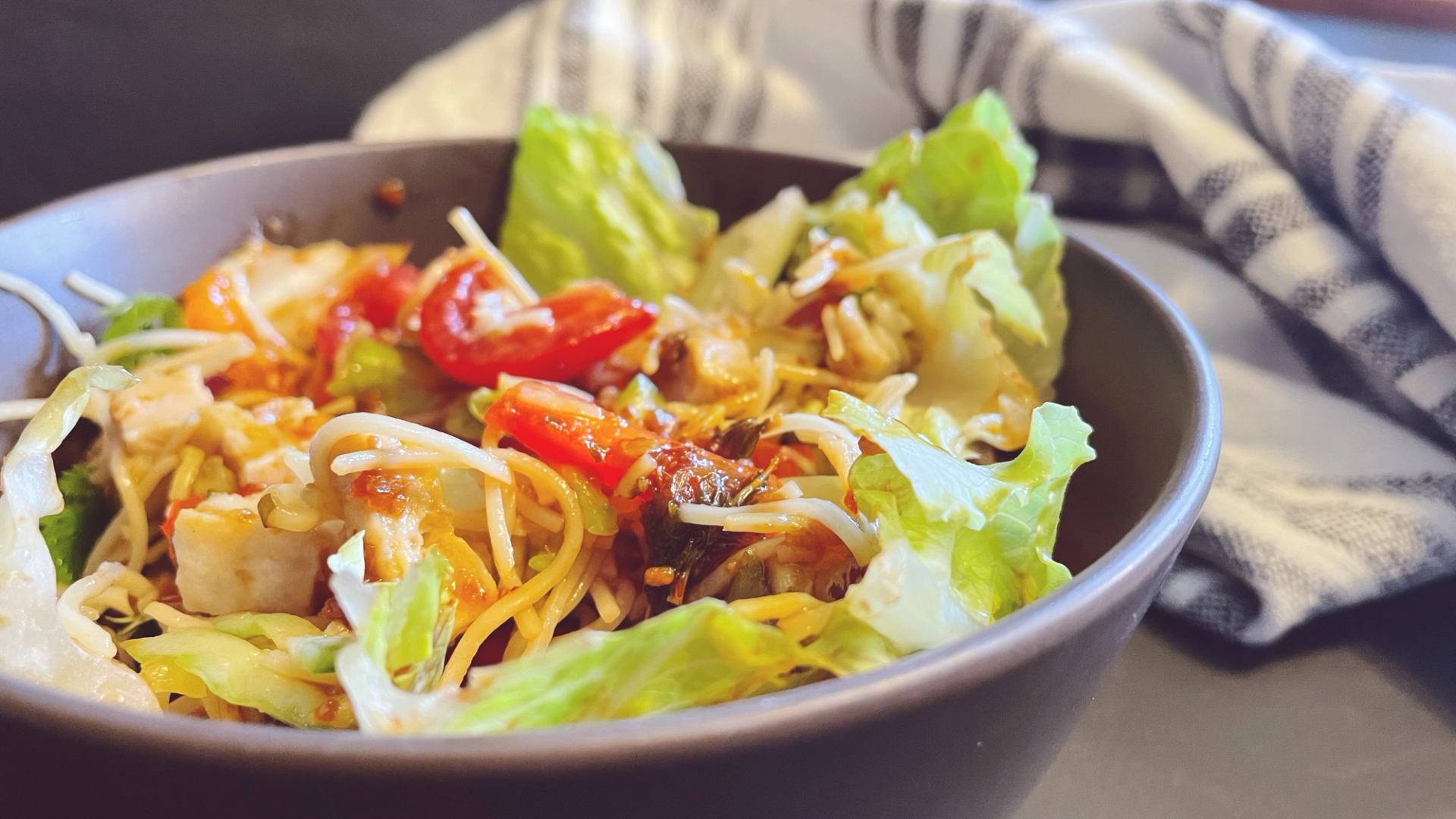 Bowl of noodle salad with lettuce, red peppers and tofu on table.