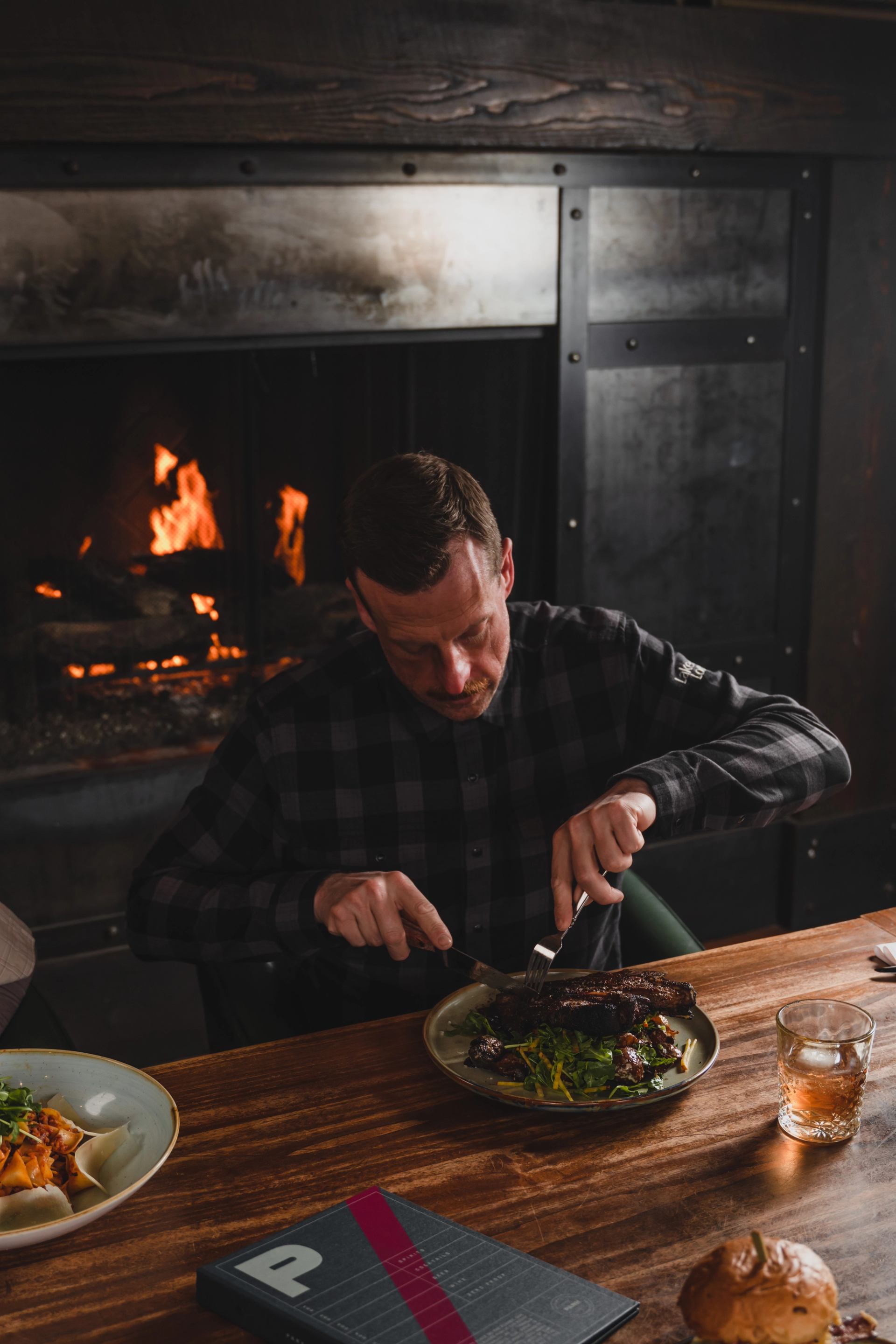 A man in a plaid shirt cuts a steak at a wooden table, with a glowing fireplace behind him.