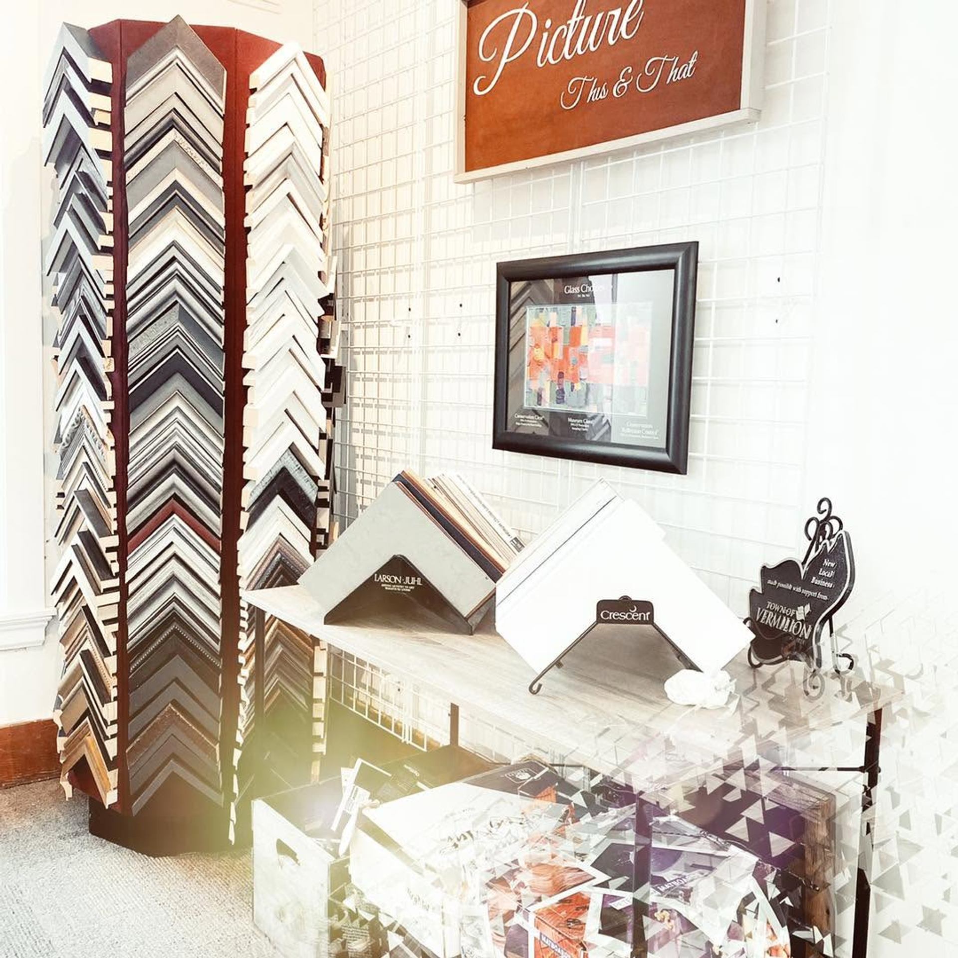 Framing shop display with corner samples, framed artwork, and matboard catalogues on shelves.