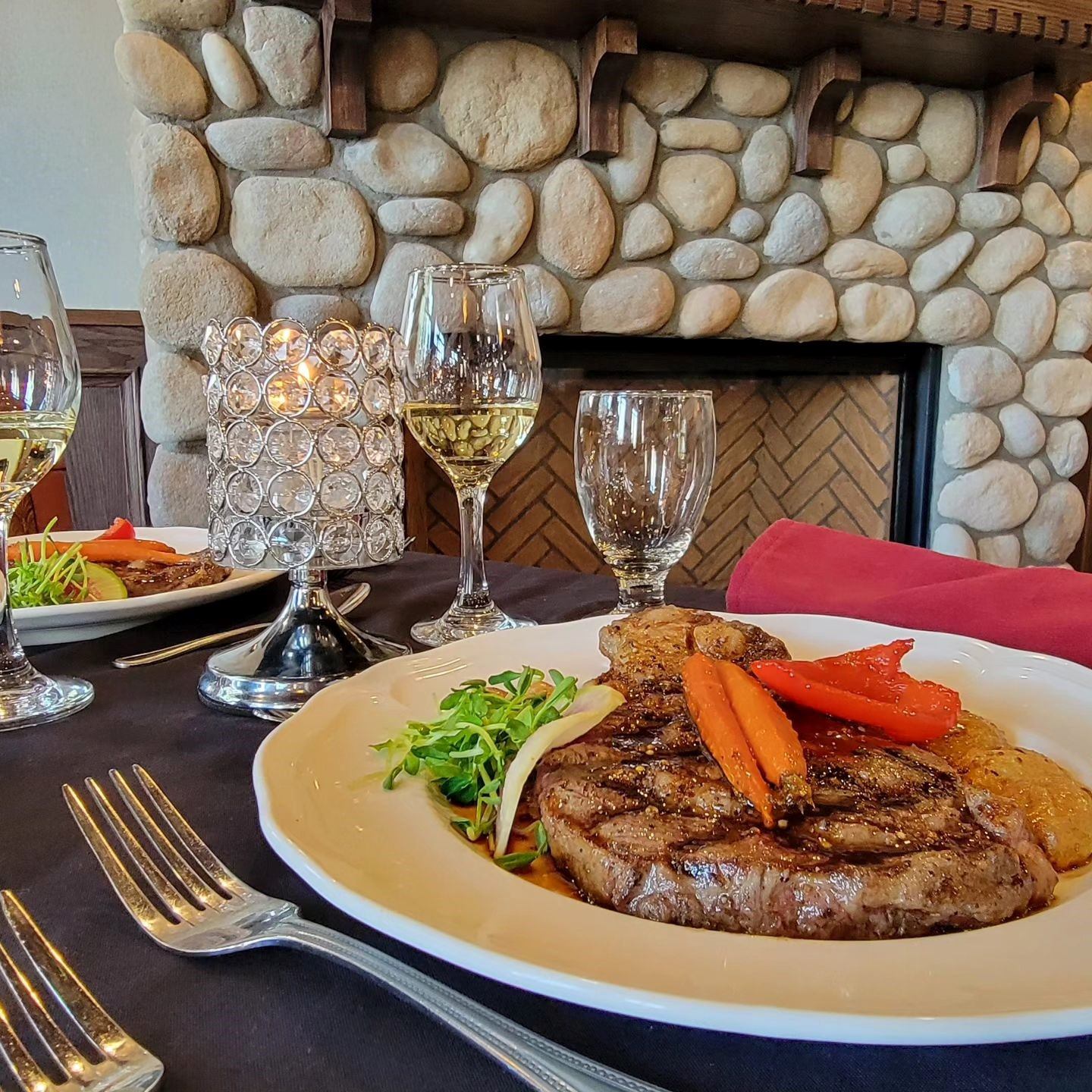 Elegant steak dinner by a stone fireplace with wine glasses and cozy ambiance.