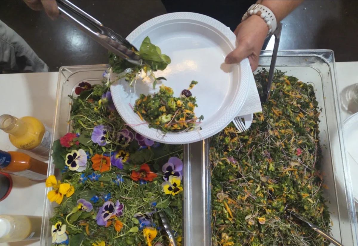 Hands serve mixed salad with edible flowers onto a white disposable plate.