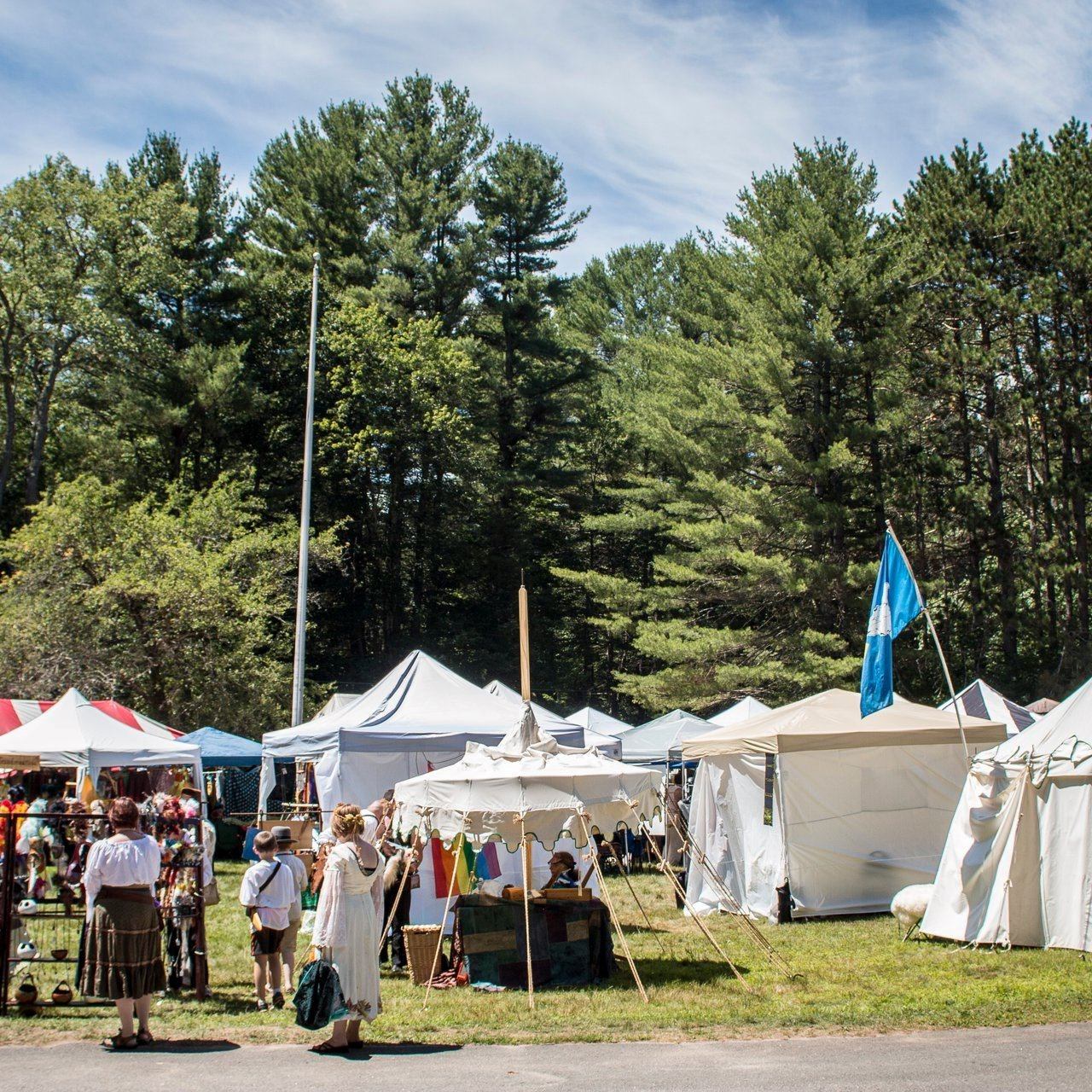 Outdoor fair with white tents, people, and a blue flag in a grassy, wooded area.