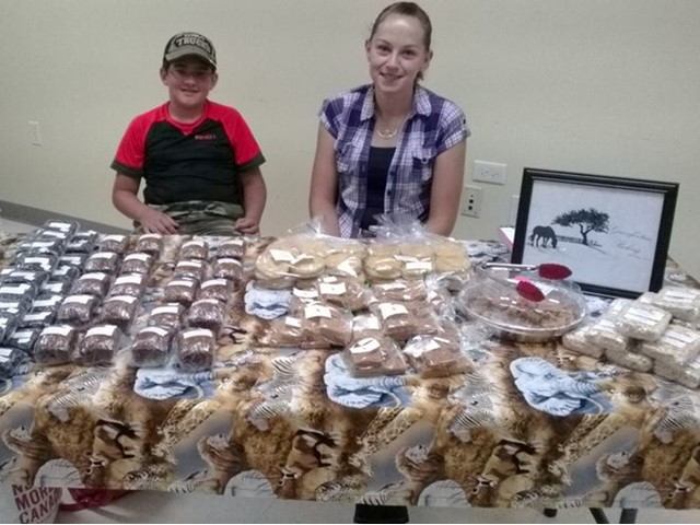 People behind a table of baked goods and framed artwork at an indoor farmers market.