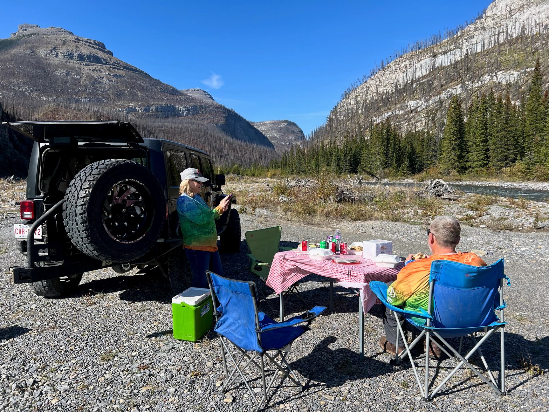 Jeep parked by a river as people relax in camping chairs with a picnic setup.