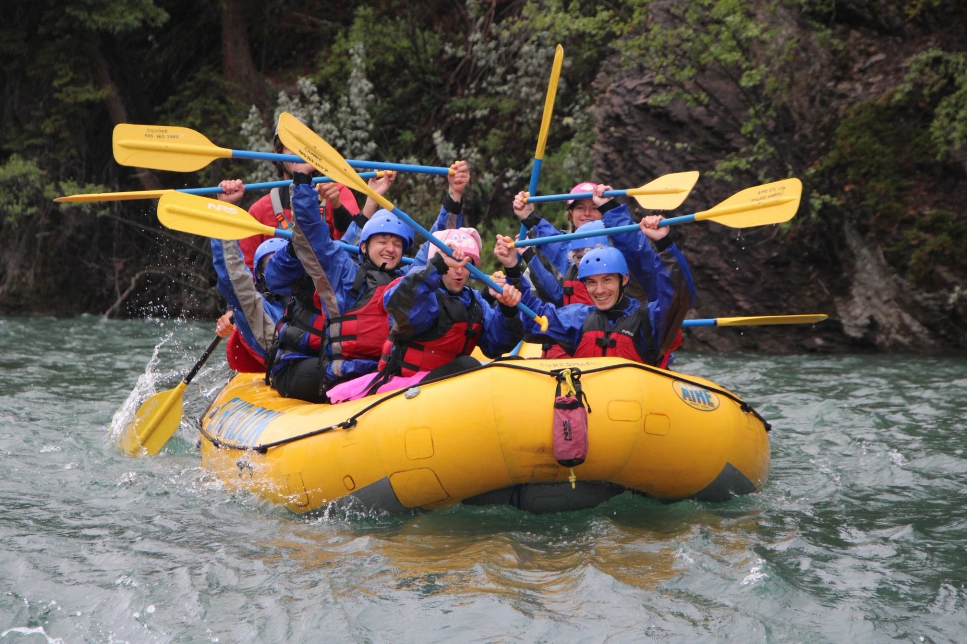 Group in helmets and lifejackets paddles yellow raft on fast river near rocky shore.