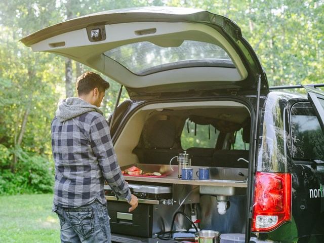 Person at the back of a campervan with a compact kitchen setup in a wooded outdoor area.