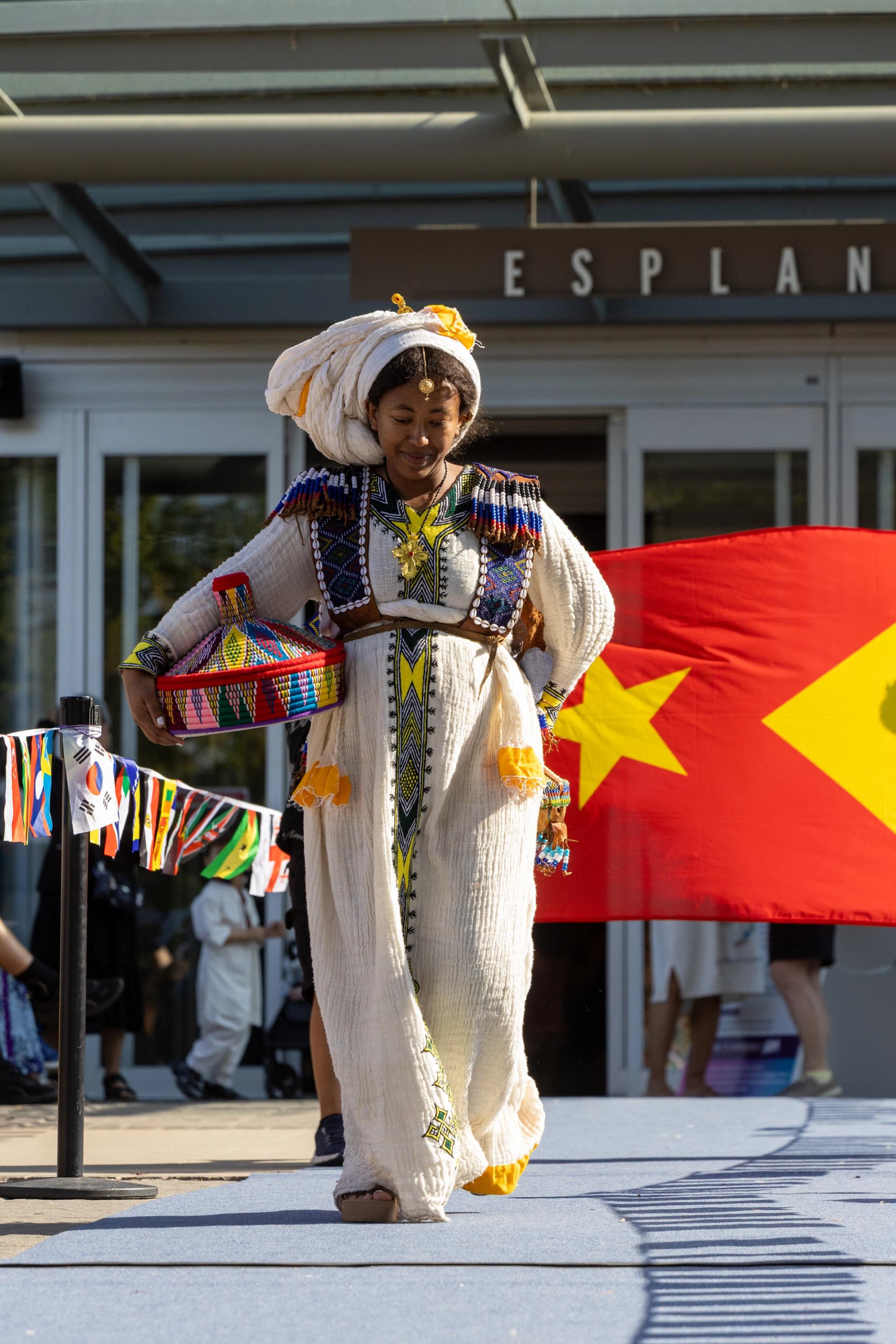 Person in traditional embroidered robe walks on runway, holding colourful basket, flags behind.