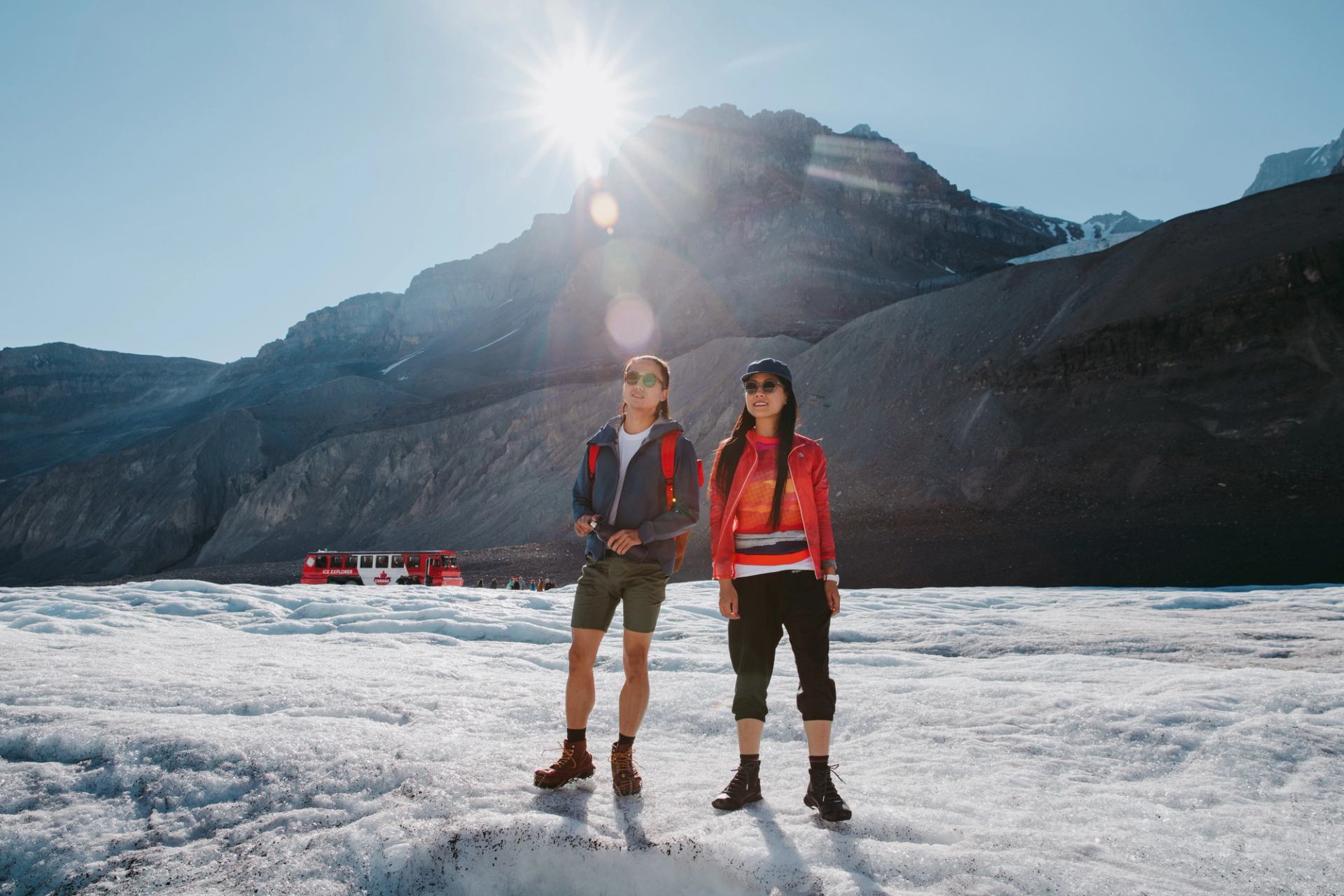 Couple standing on the Columbia Glacier with a red Ice Explorer bus in the background.