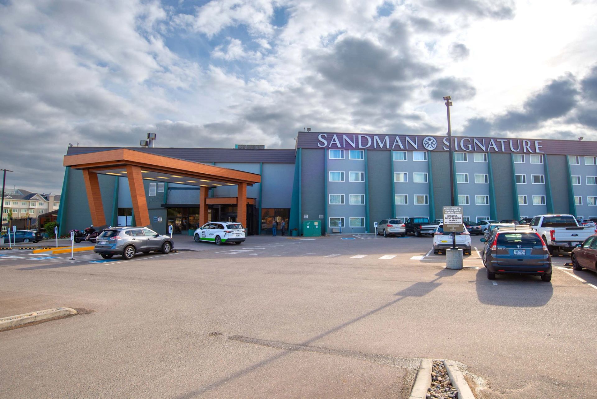 Sandman Signature hotel exterior with covered entrance, parked cars and cloudy sky above..