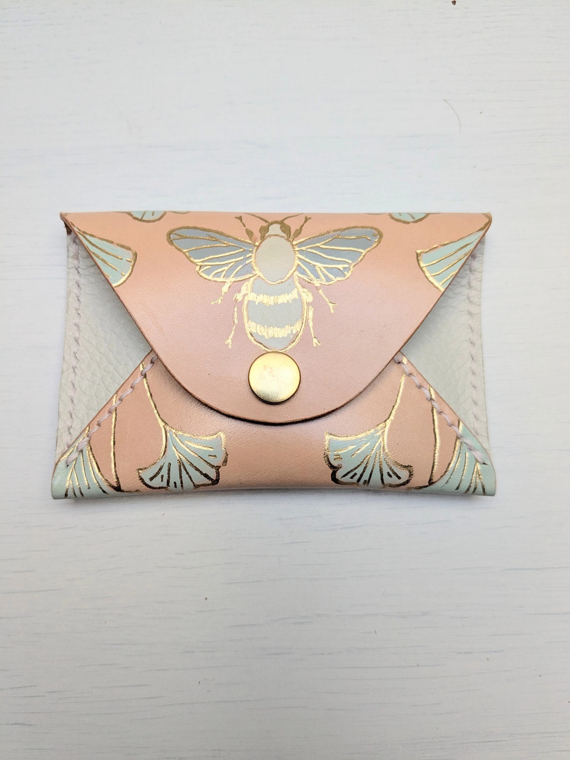 A peach and white leather card holder featuring a gold bee and light blue ginkgo leaf design, fastened with a gold snap.